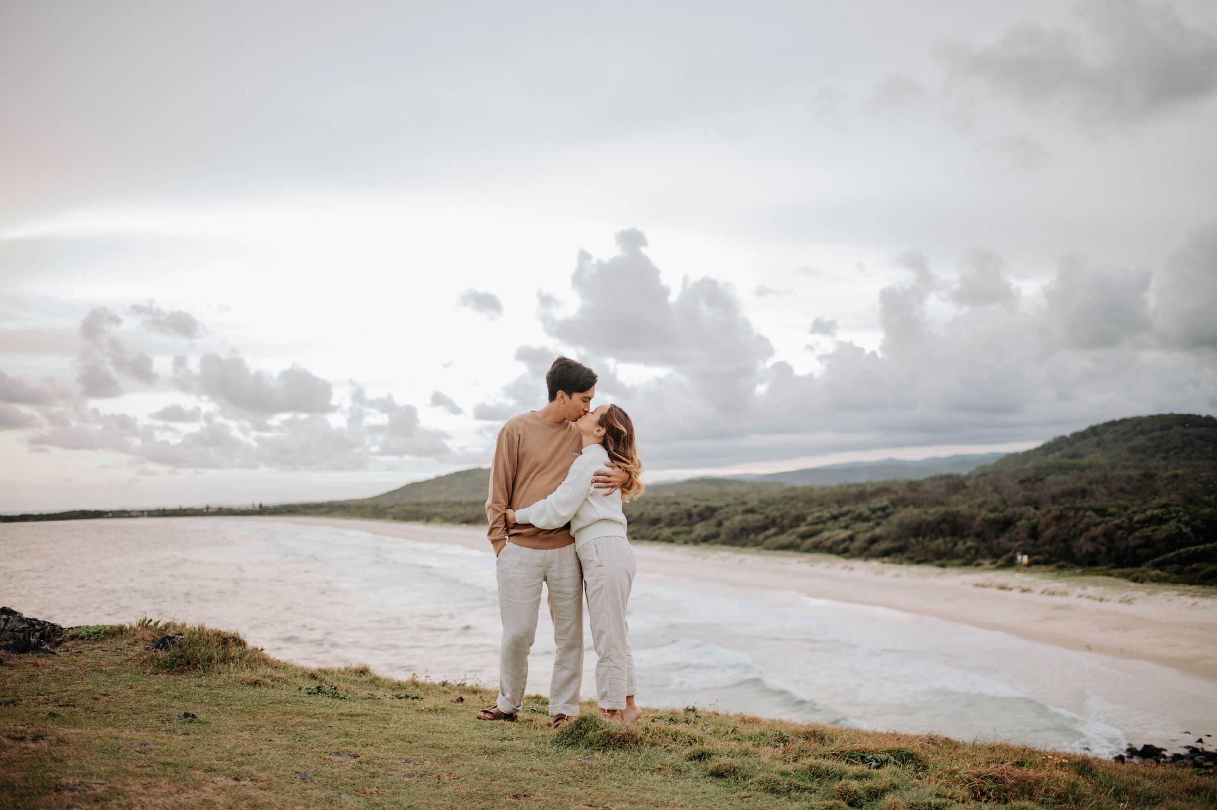 A couple in white outfits shares an intimate moment overlooking a coastal landscape at sunset.