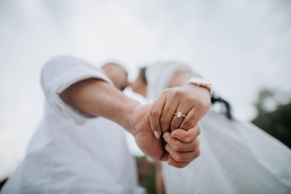Close up of two hands clasped together showing off an engagement ring against a blurred background.