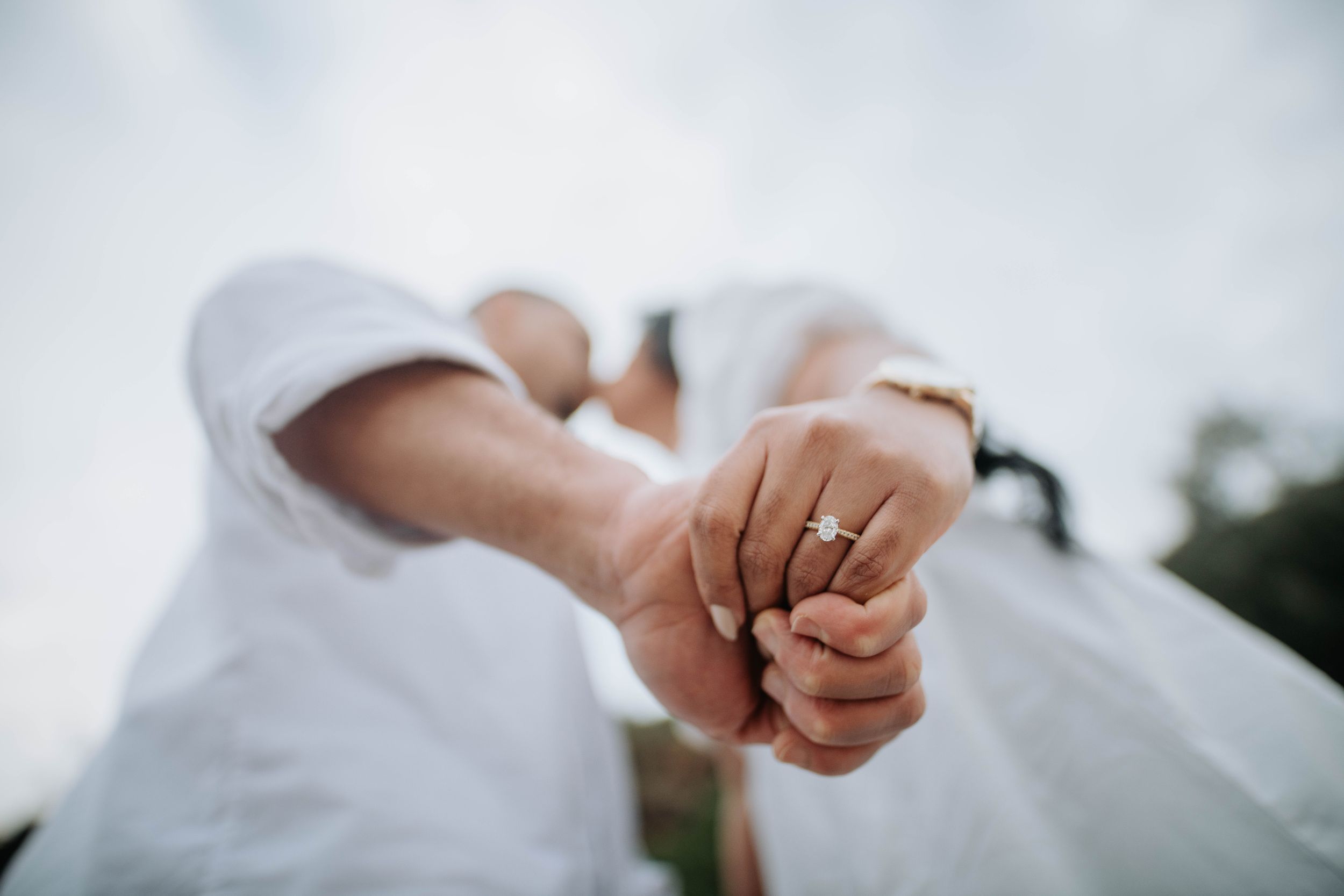Close up of two hands clasped together showing off an engagement ring against a blurred background.
