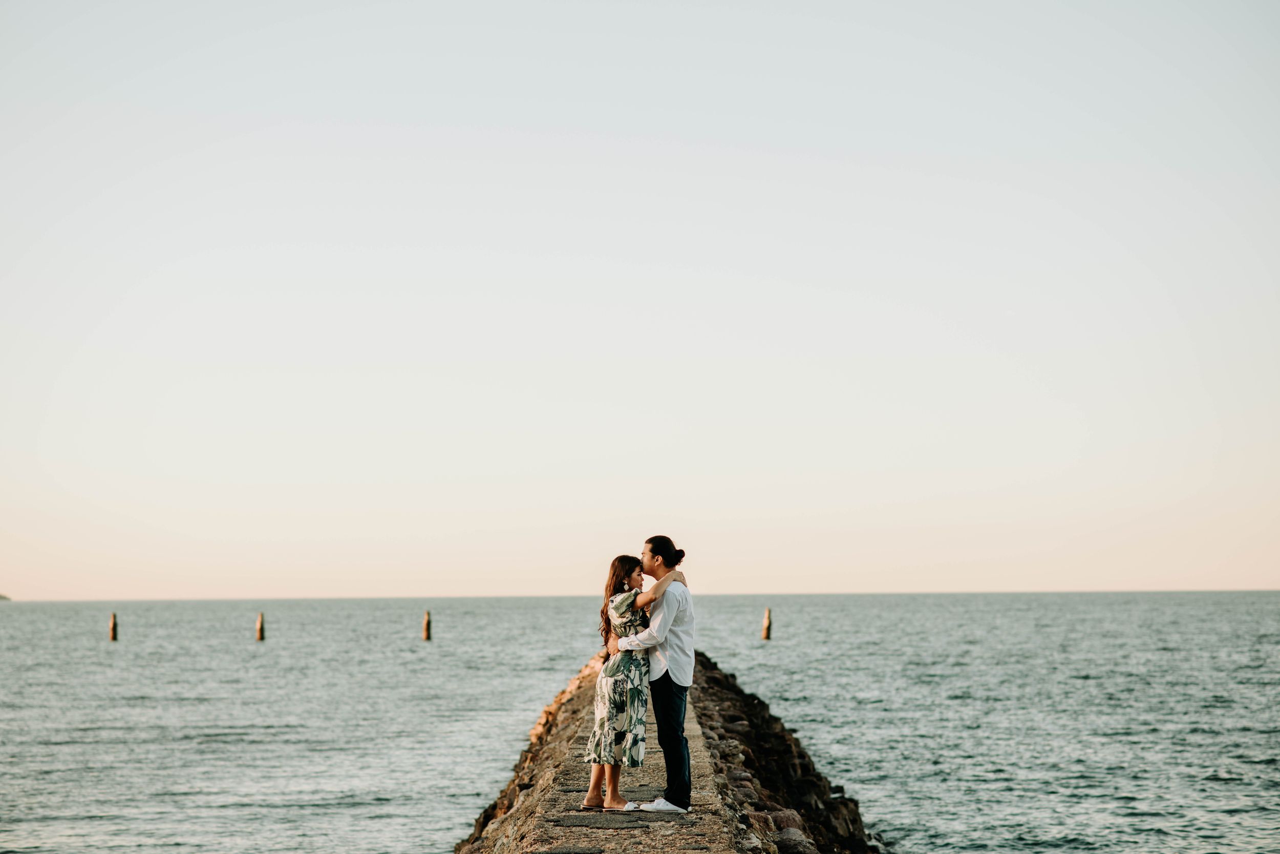 A couple shares a romantic moment on a stone jetty extending into the ocean at sunset.