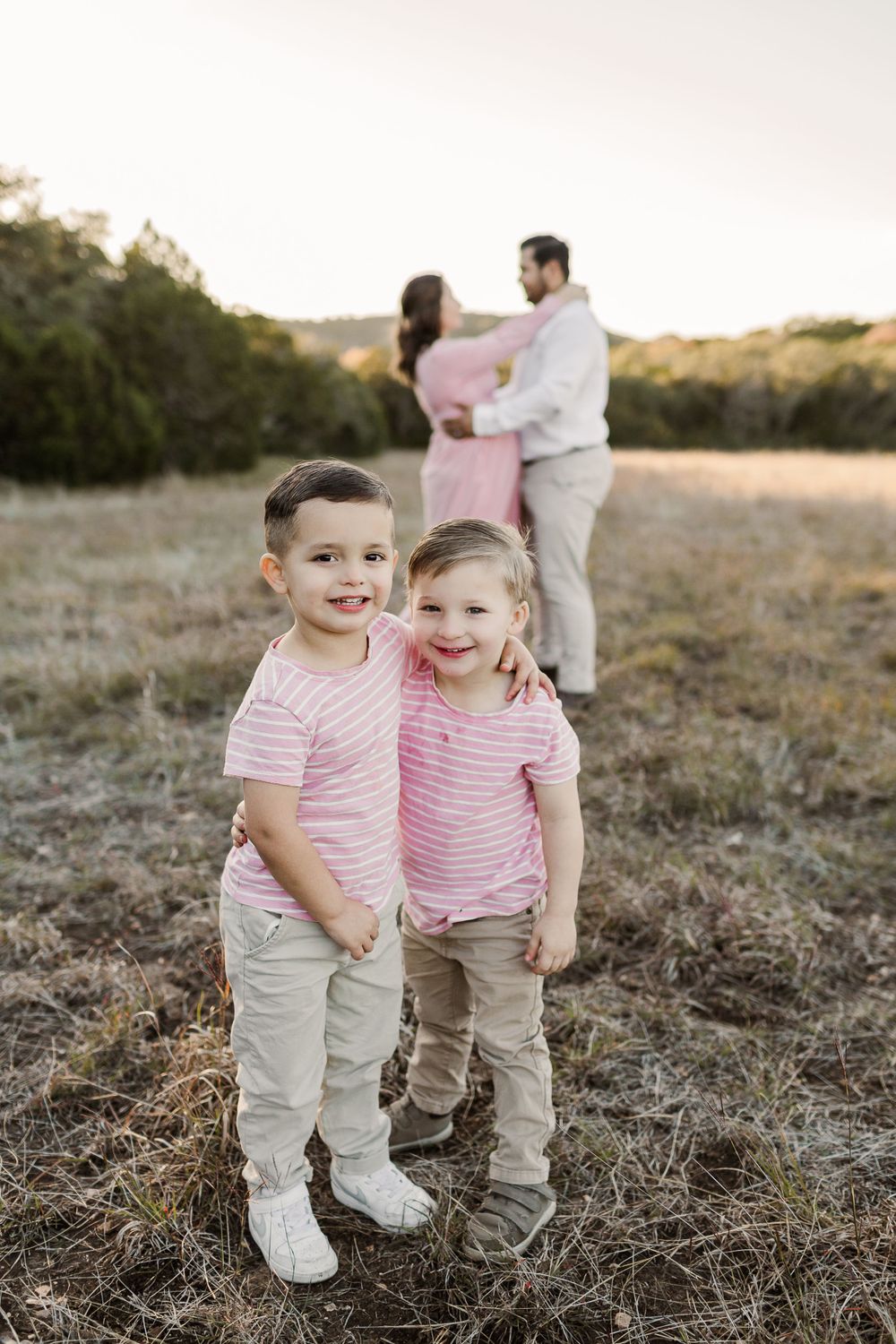 Two children in matching pink striped shirts pose while parents embrace in background.