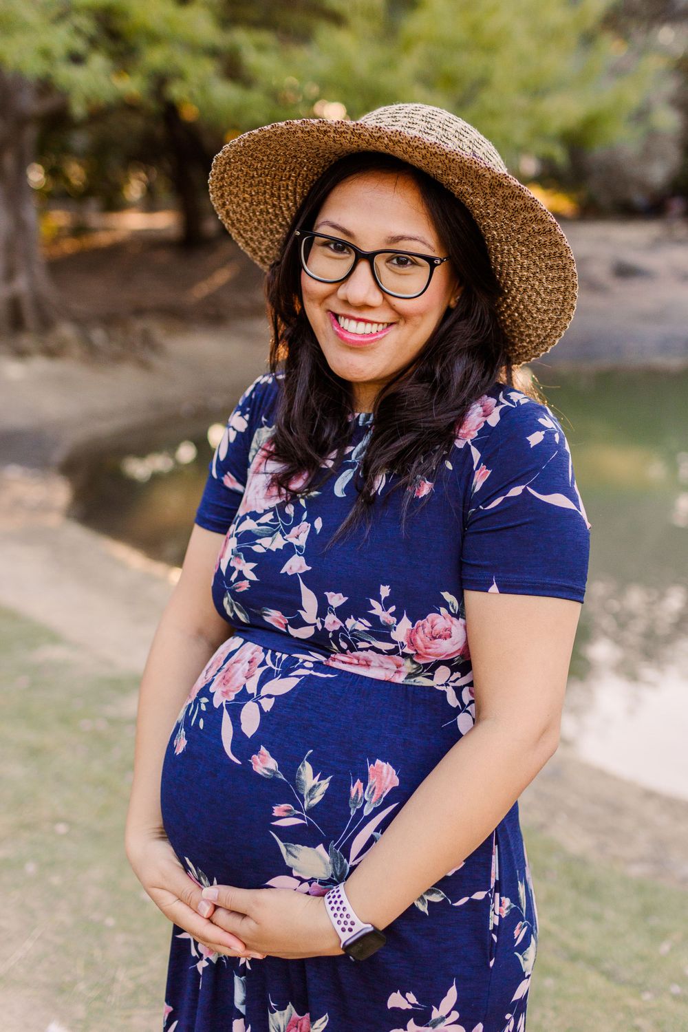 Maternity portrait in blue floral dress and straw hat by waterfront.
