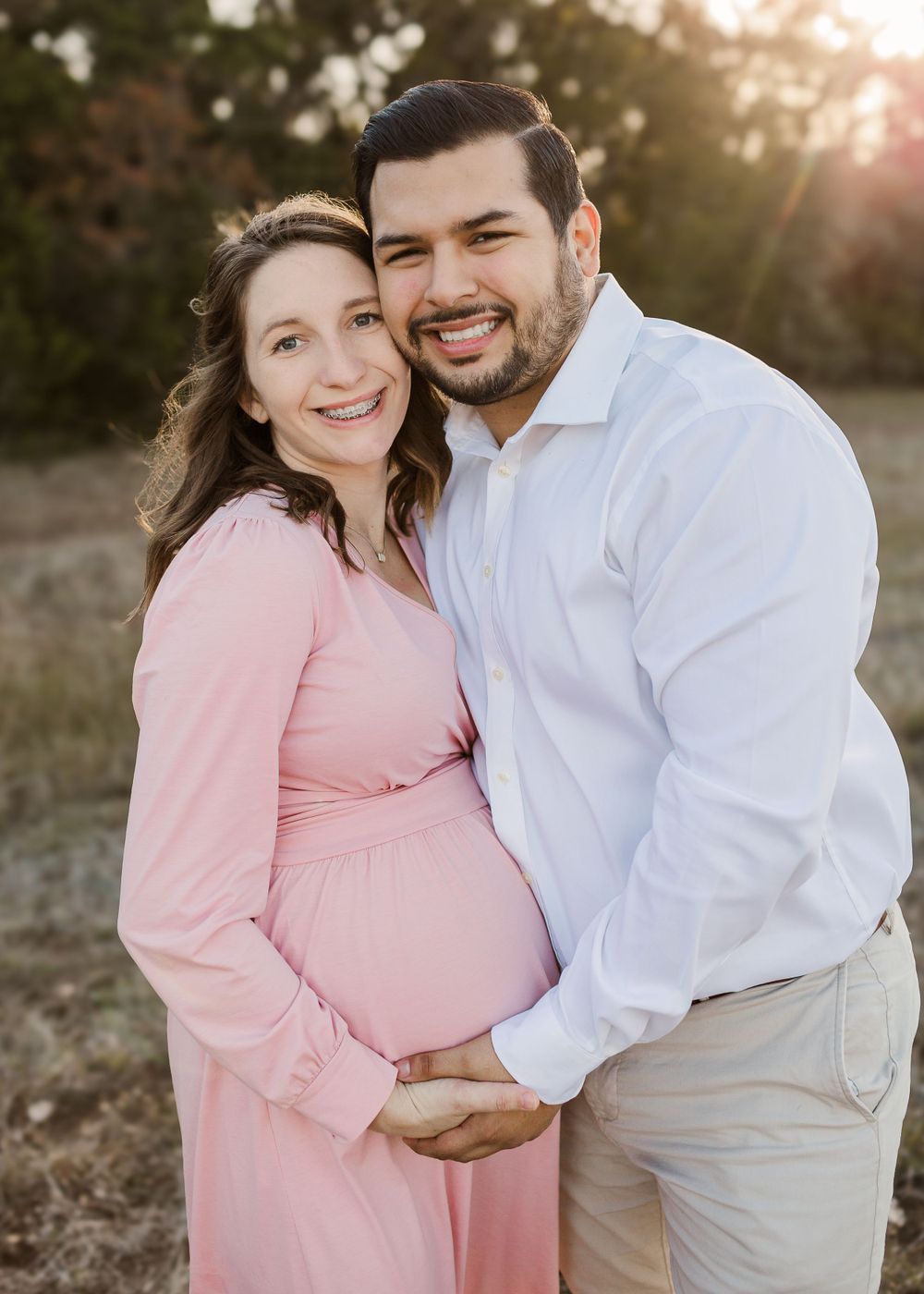 Couple embracing outdoors with woman in pink maternity dress during sunset.