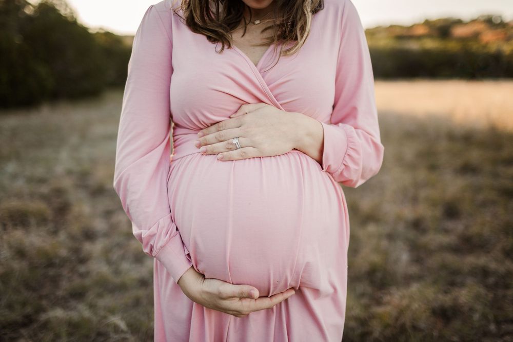 Close-up of pregnant belly with hands tenderly placed on pink maternity dress.