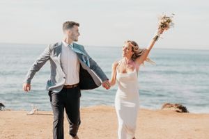 Joyful couple celebrating on coastal cliff with bouquet raised in celebration.