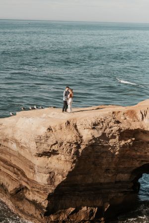Aerial view of couple standing on dramatic ocean cliff during sunset ceremony.