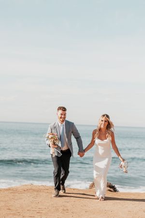 Couple holding hands while walking along beach cliff carrying wedding bouquets.