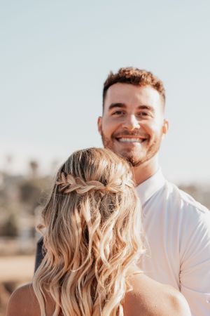 A romantic photo of a couple in warm sunlight, the man smiling while the woman's blonde hair is styled with braids.