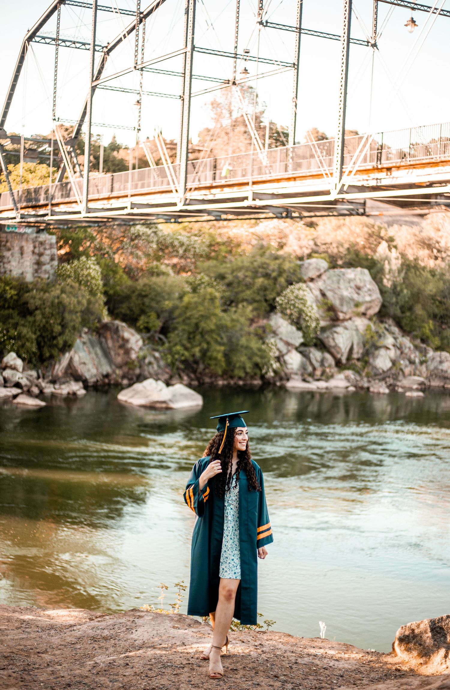A graduate poses by a steel bridge over scenic rocky waters wearing traditional graduation regalia.