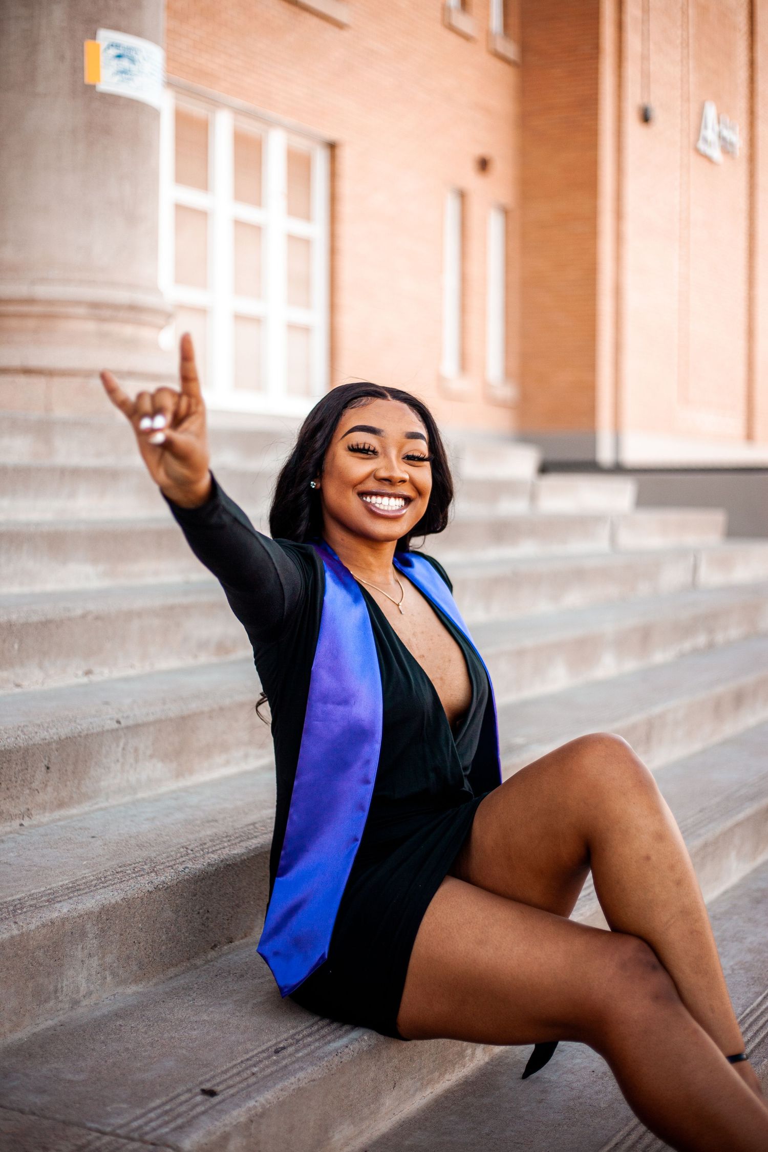 Graduate in graduation attire sitting on stone steps making celebratory gesture.