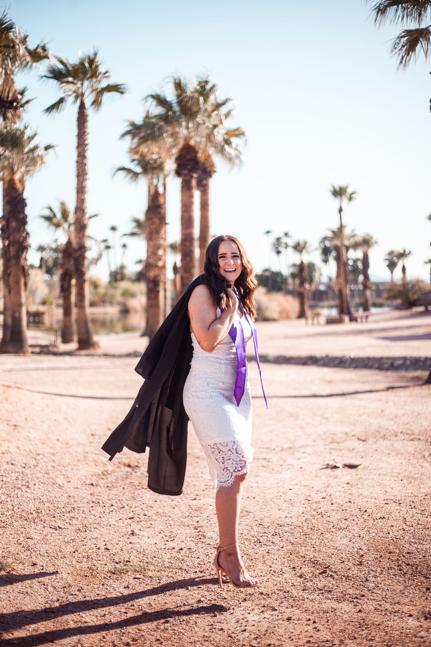 Graduate in white dress and purple stole posing among palm trees in desert setting.