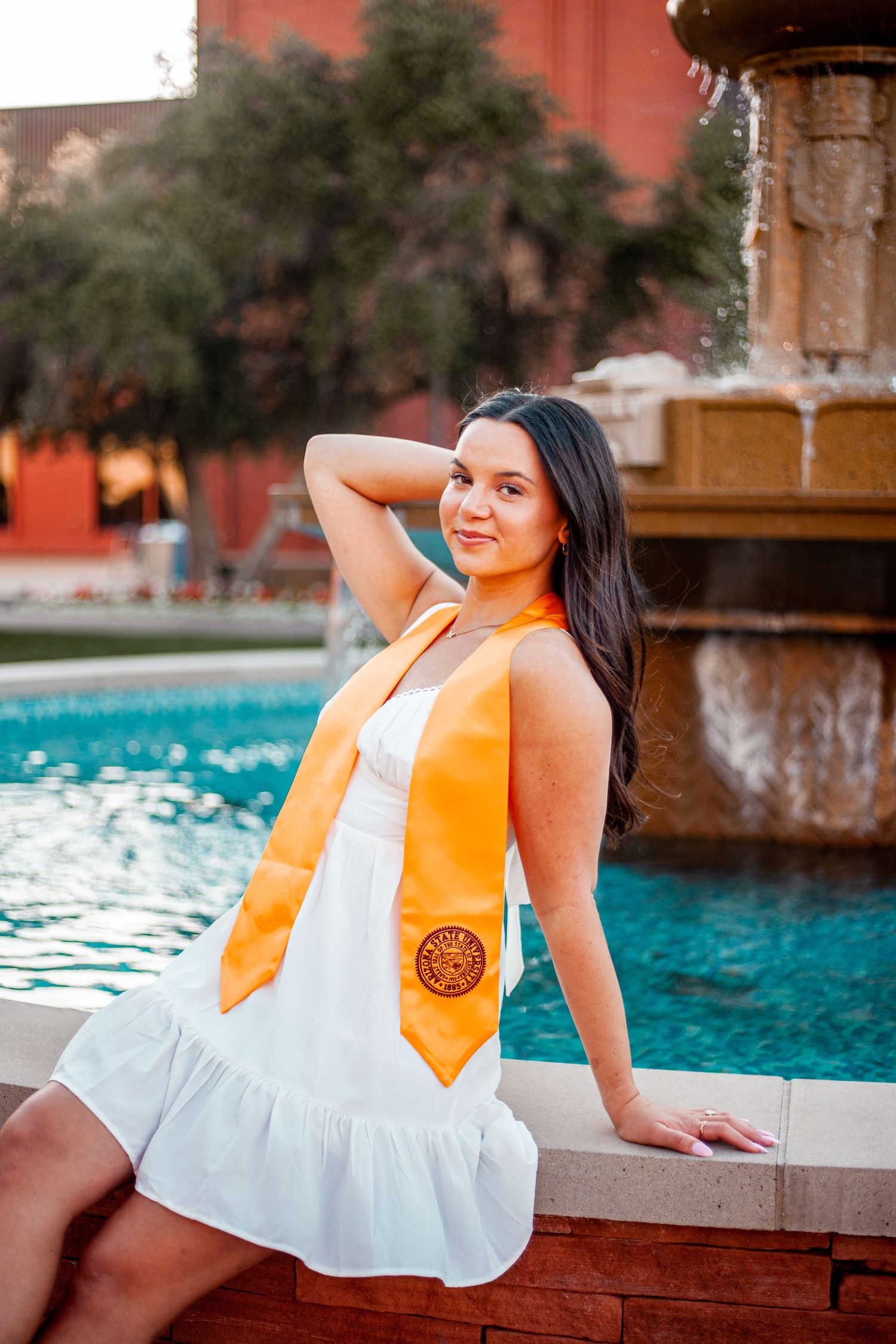 A festive graduation portrait by a fountain featuring a white dress and orange stole.