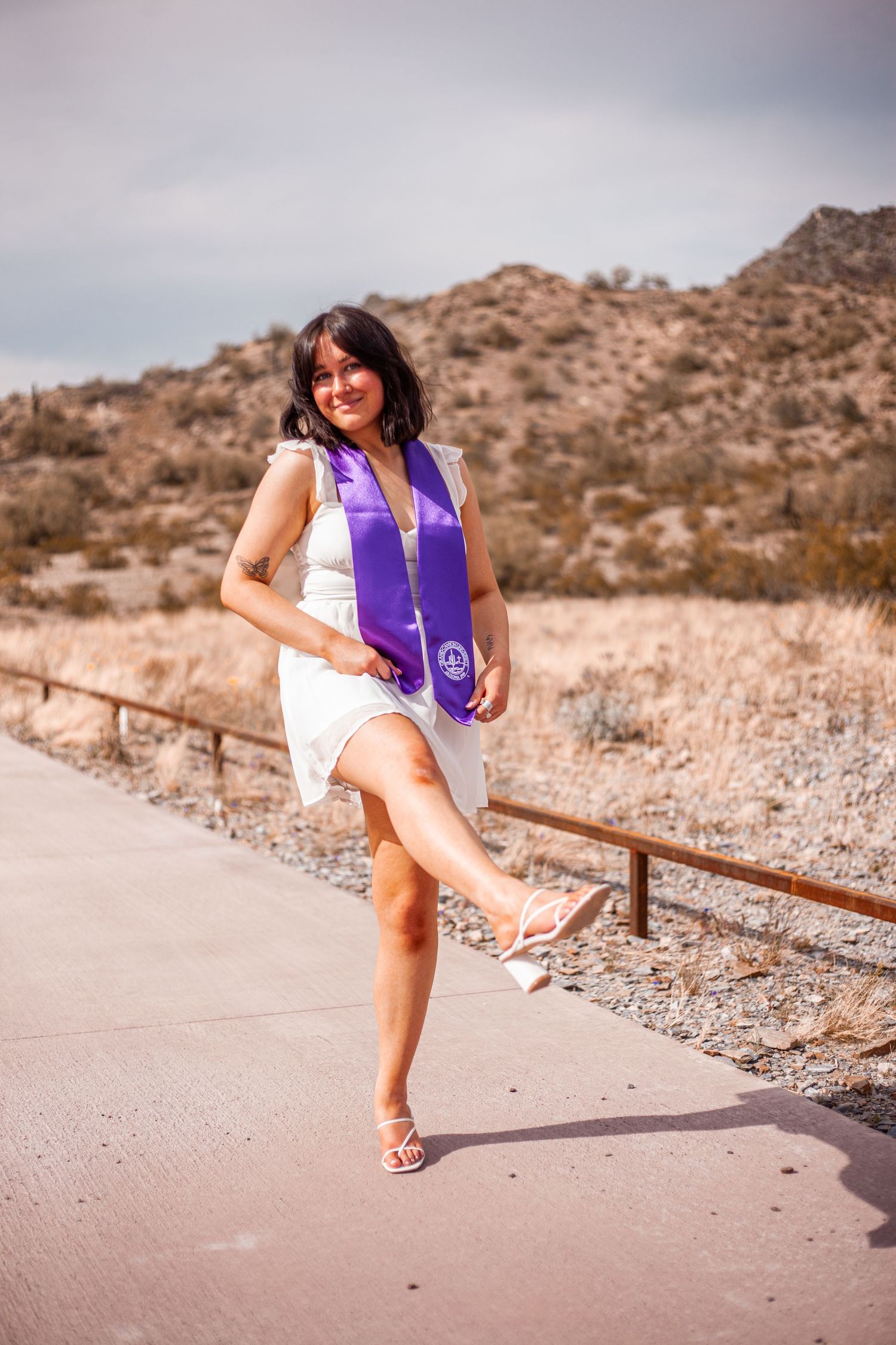 Graduate in white dress and purple stole posing on desert path with mountains in background.