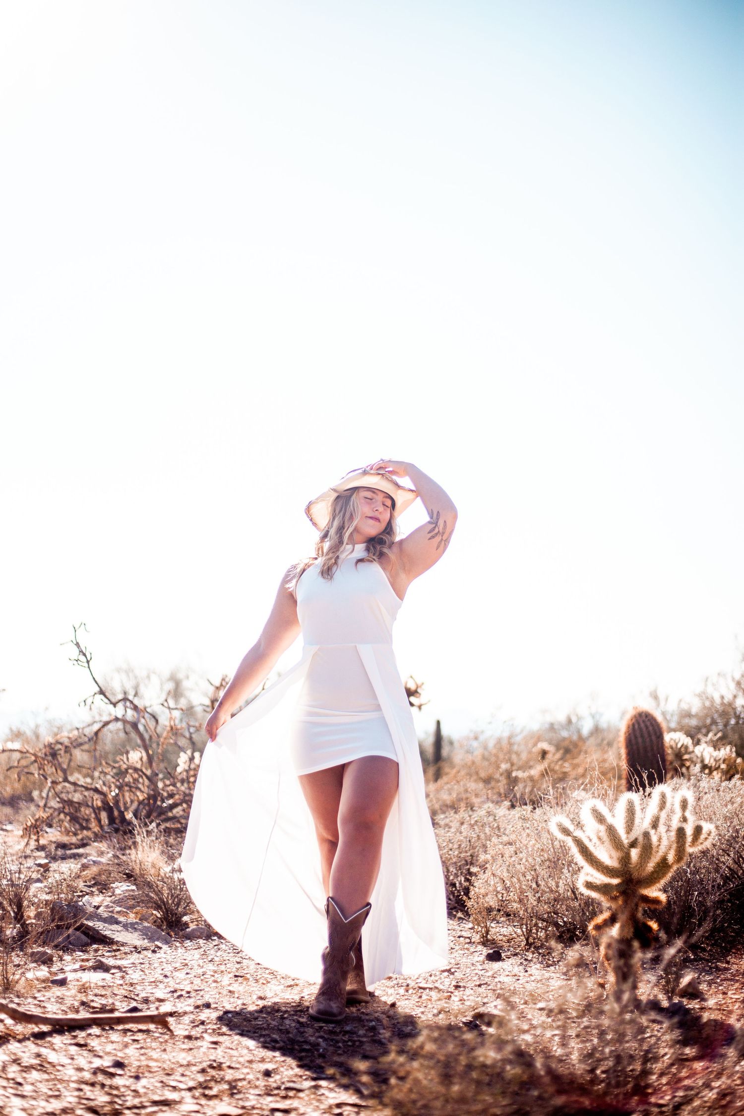 Figure in white dress and hat poses dramatically in desert landscape with cacti.