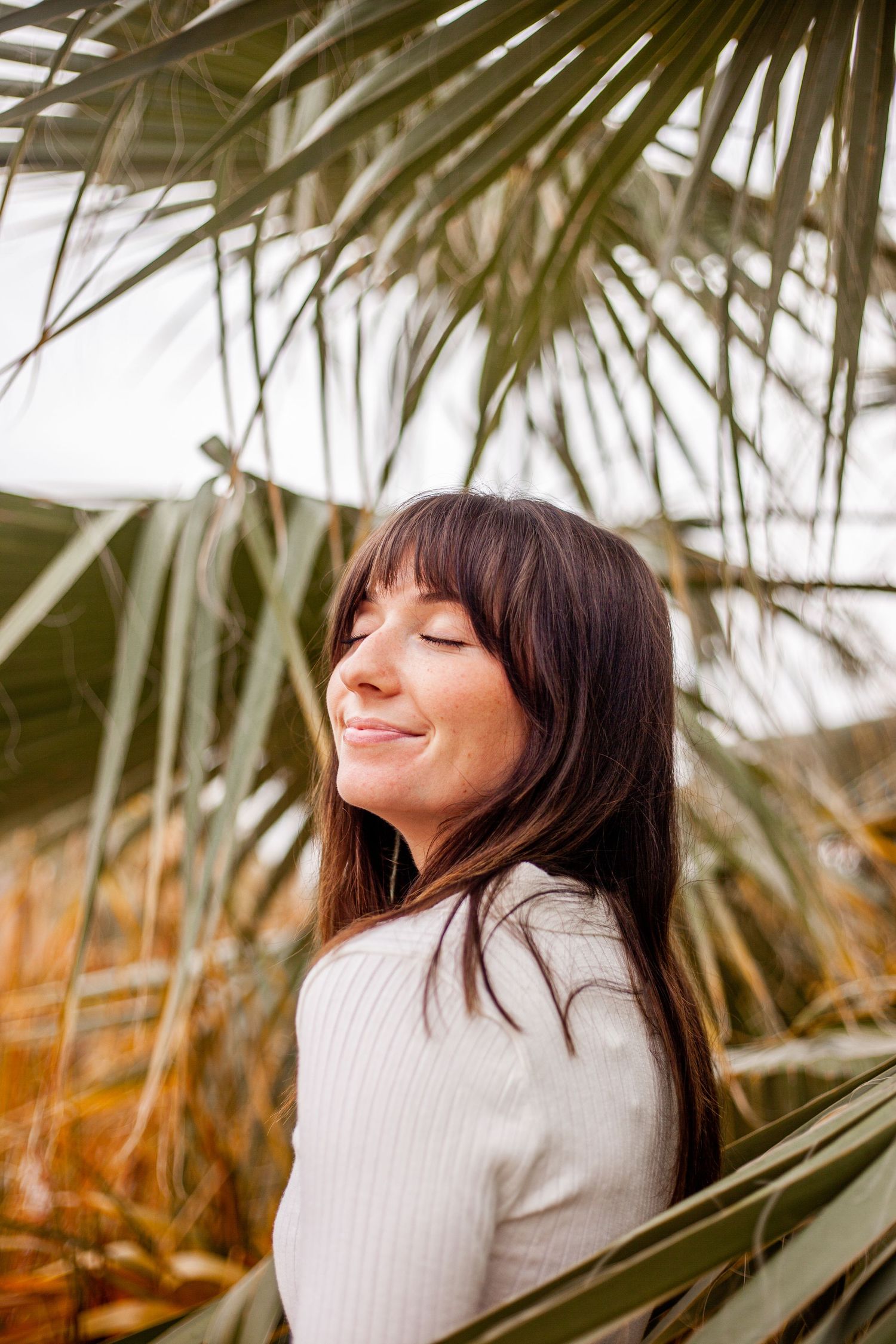 A serene moment captured with palm fronds framing a relaxed pose in a cream sweater against a bright background.