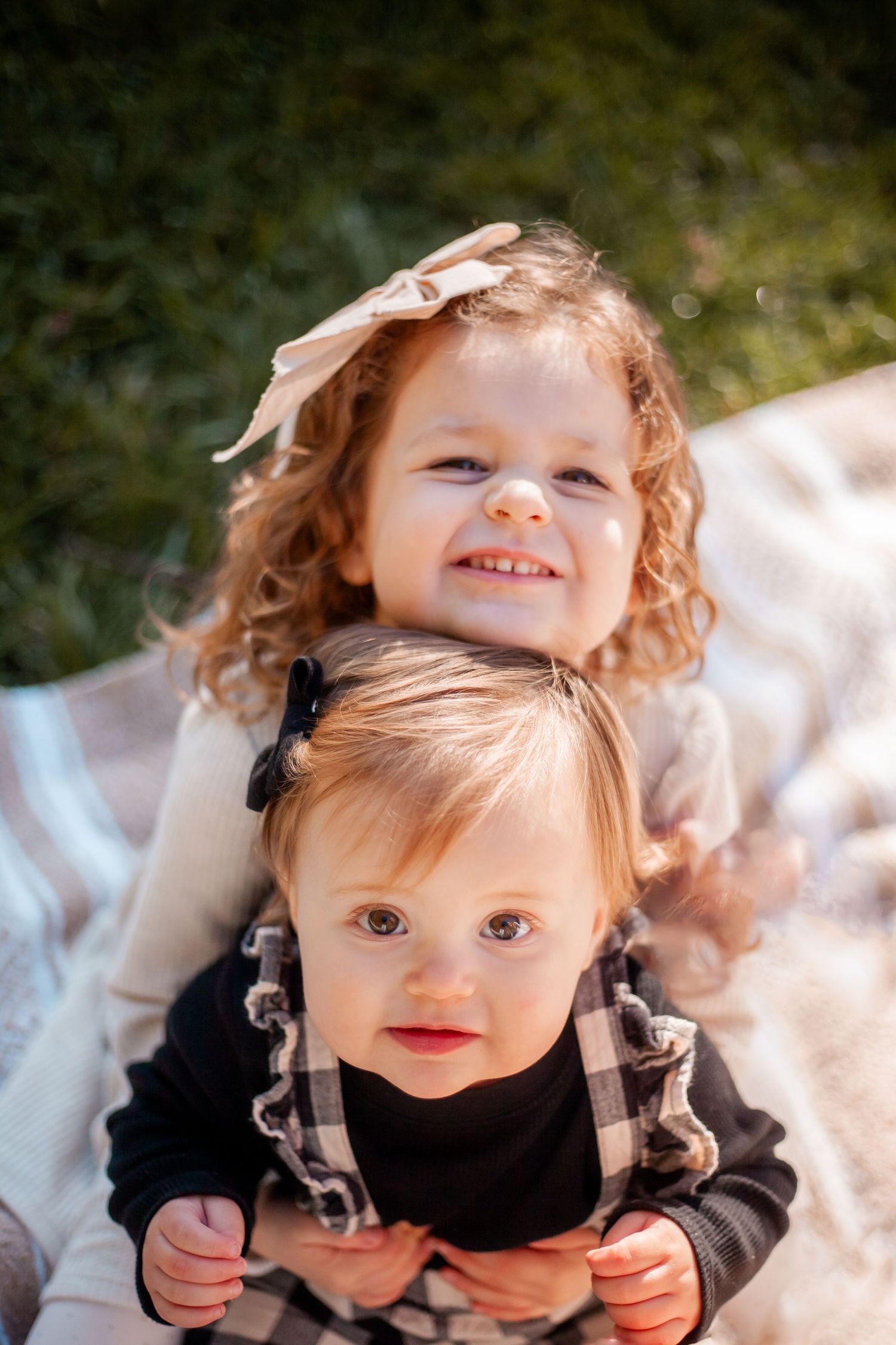 Two young children in light-colored outfits sitting together on blanket outdoors.