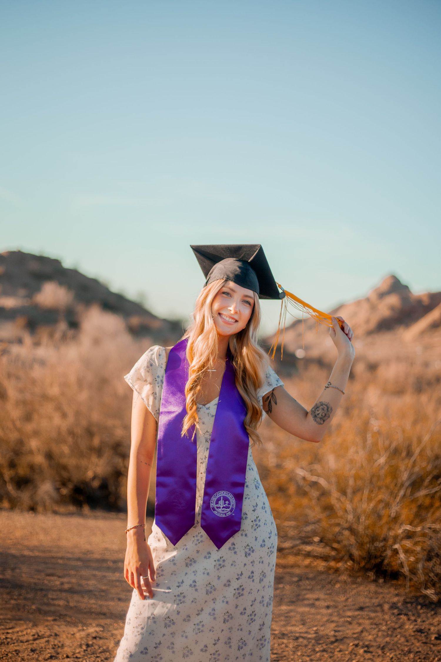 Graduate in purple stole and cap poses in desert landscape at sunset.