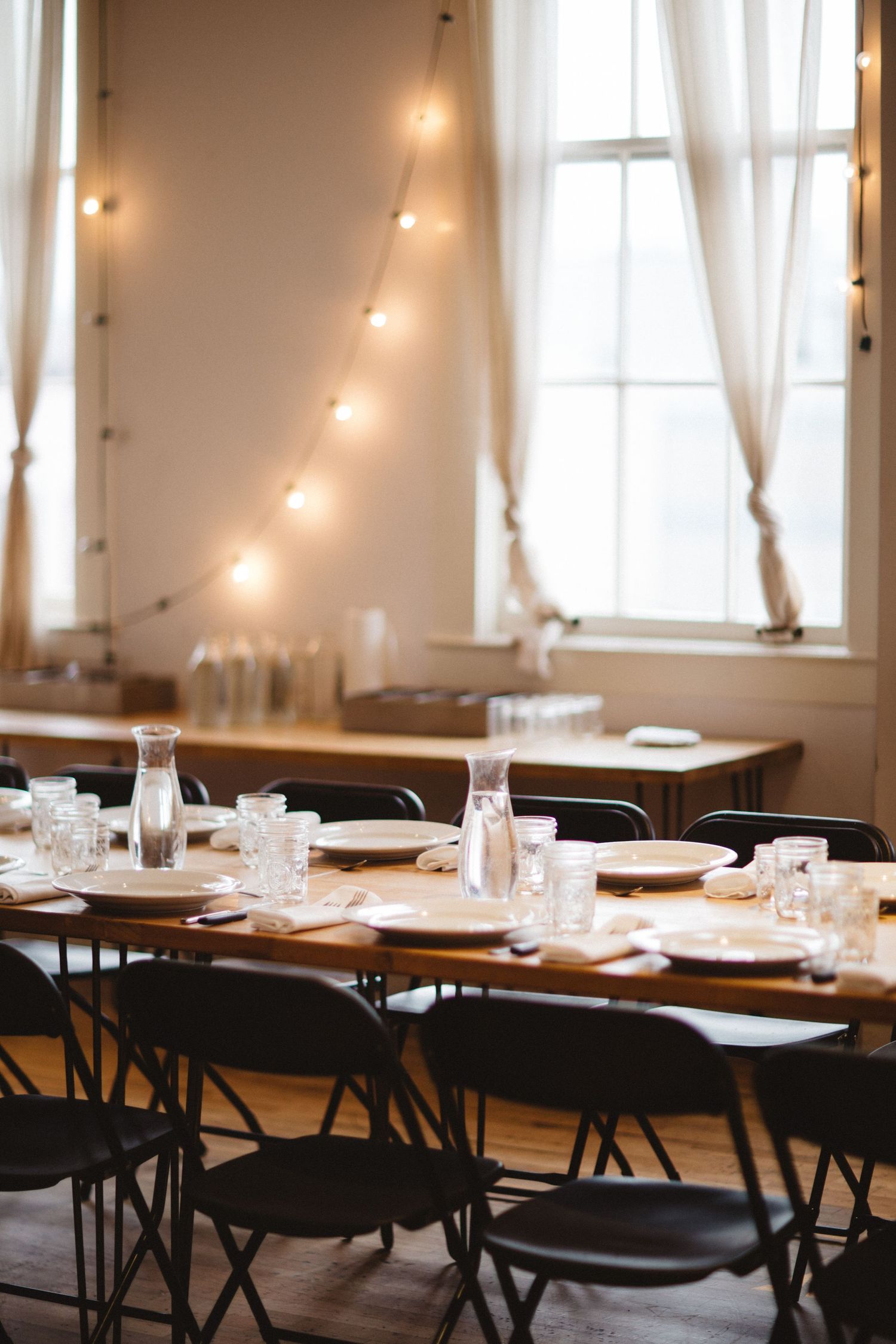An elegant dining setup with string lights and table settings in a bright room with tall windows.