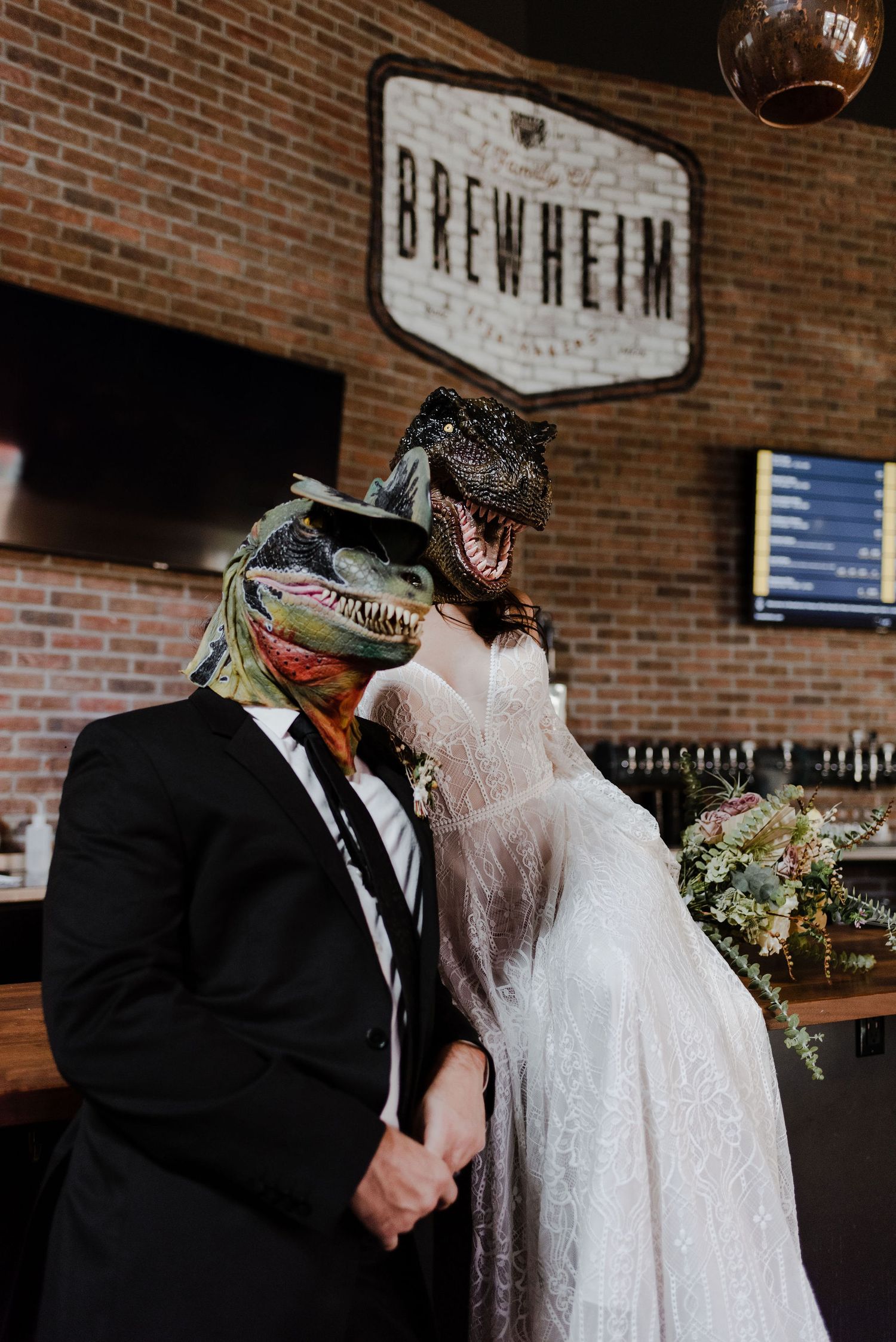 A couple in wedding attire sit on the bar of a small Anaheim brewery posing wearing dinosaur masks.
