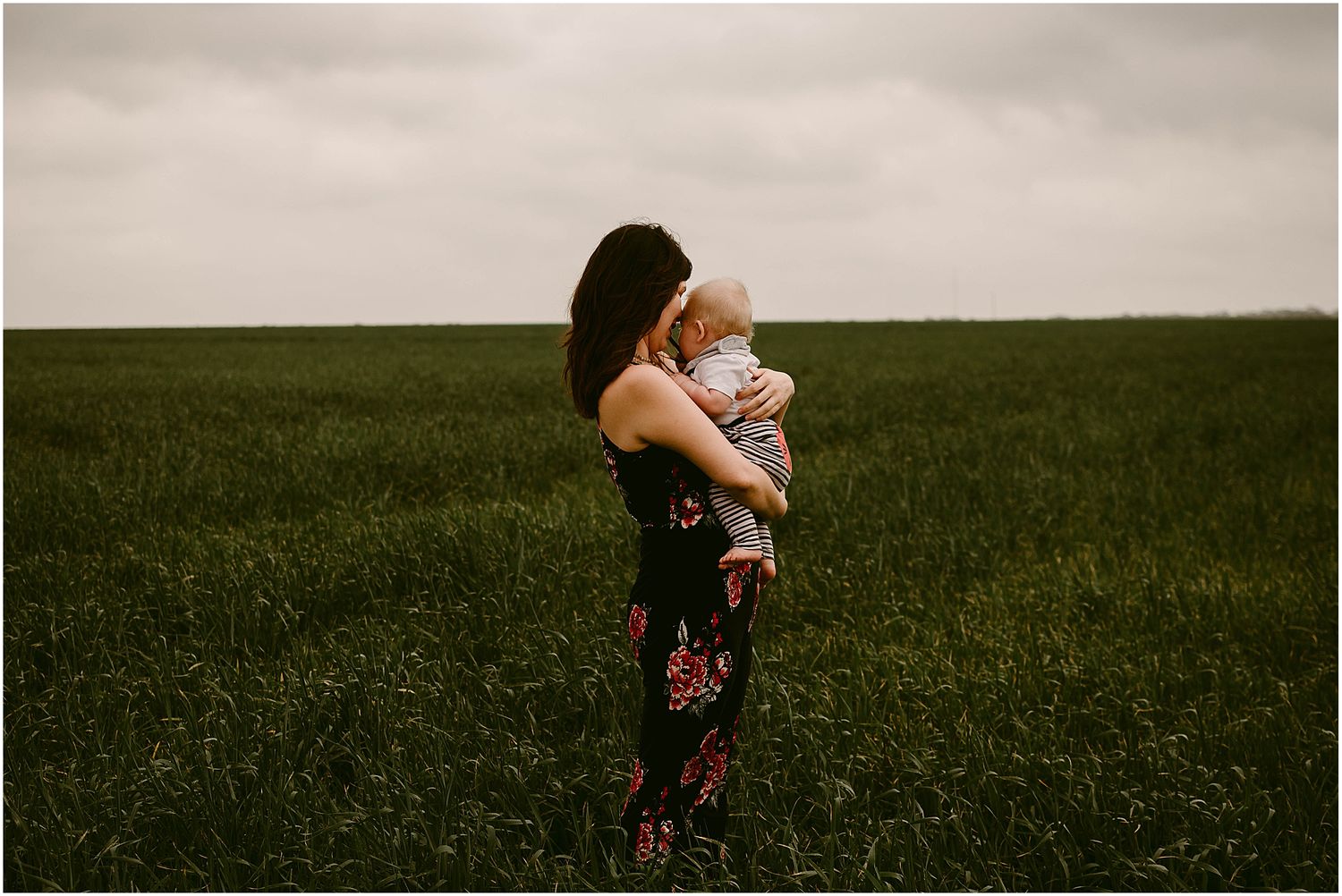 Moody portrait of adult and baby standing together in an expansive green field.