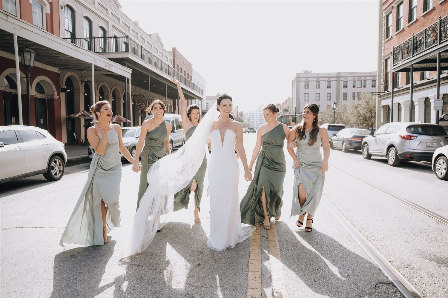 A bridal party in light colored dresses walks down an urban street on a sunny day.