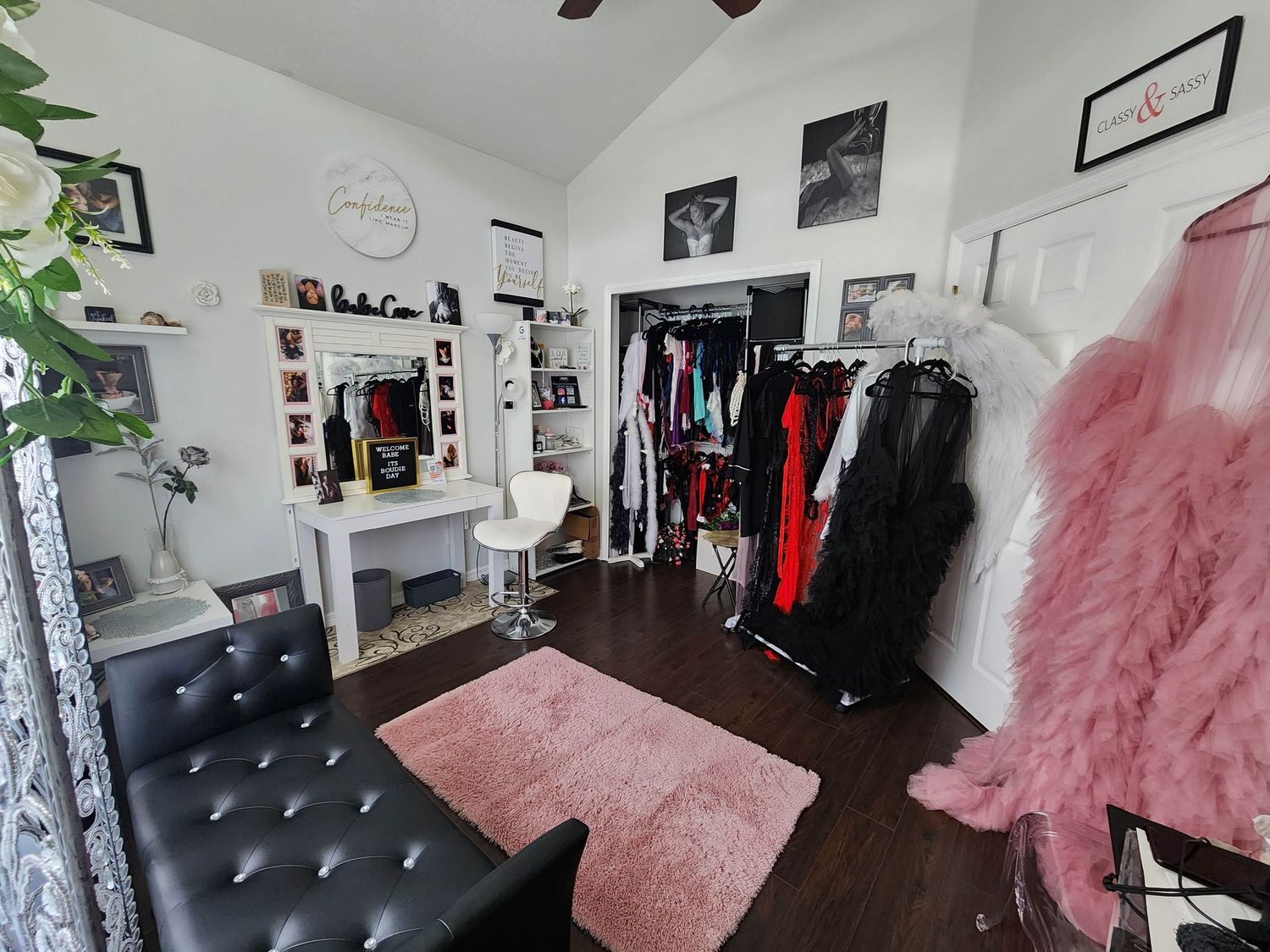 A cozy dressing room with black tufted seating, pink decor, clothing racks, and a vanity mirror setup.