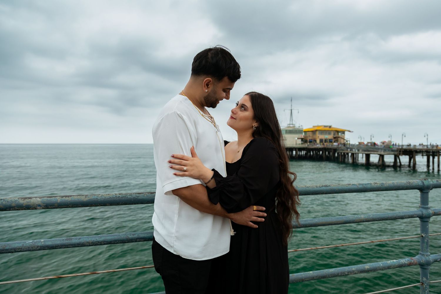 A romantic couple embracing on a pier overlooking the ocean with cloudy skies.