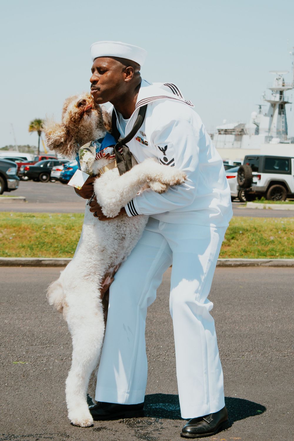 Service member in white uniform kneels to embrace fluffy white dog during homecoming.