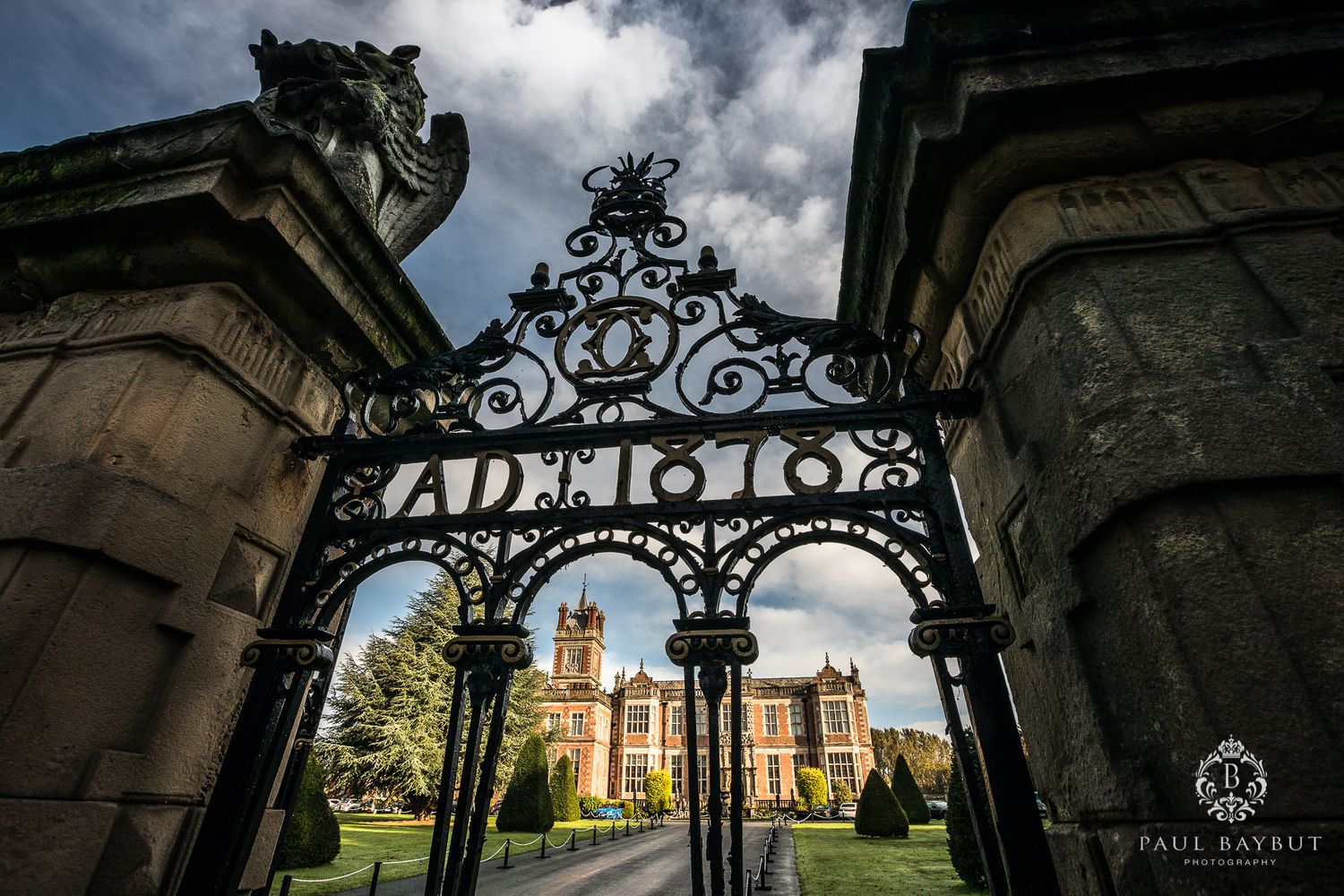 Ornate iron entrance gates to historic manor with Gothic architecture visible beyond.