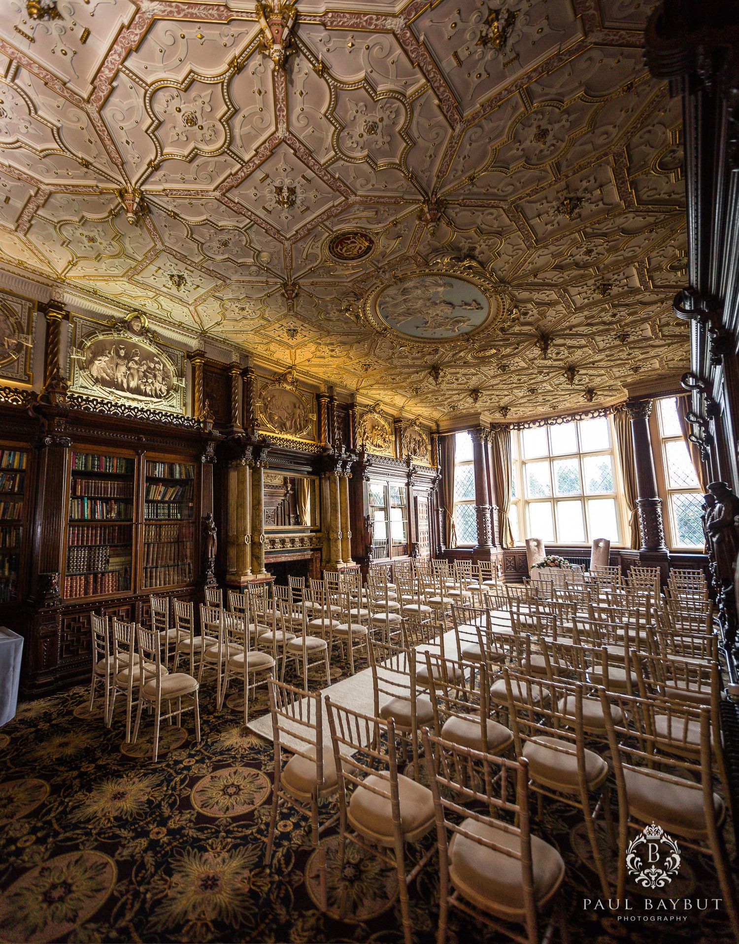 Elegant room with ornate ceiling, library shelves and rows of white chairs set up for an event.