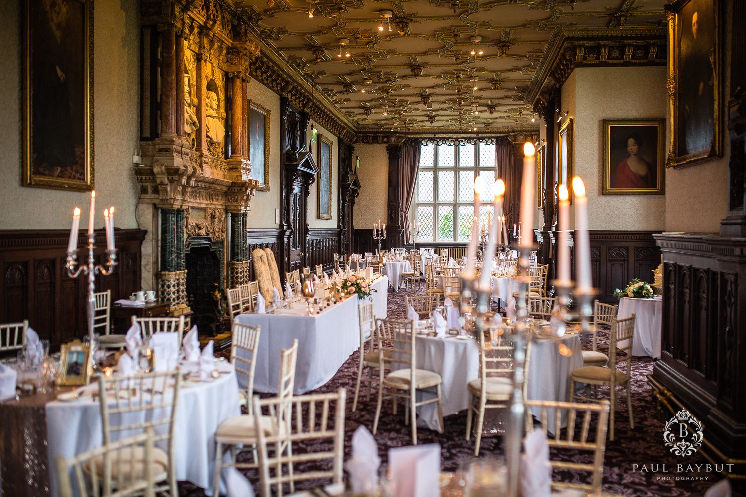 Elegant wedding reception venue with ornate fireplace, large windows, and tables set with white linens and candles in a historic hall.