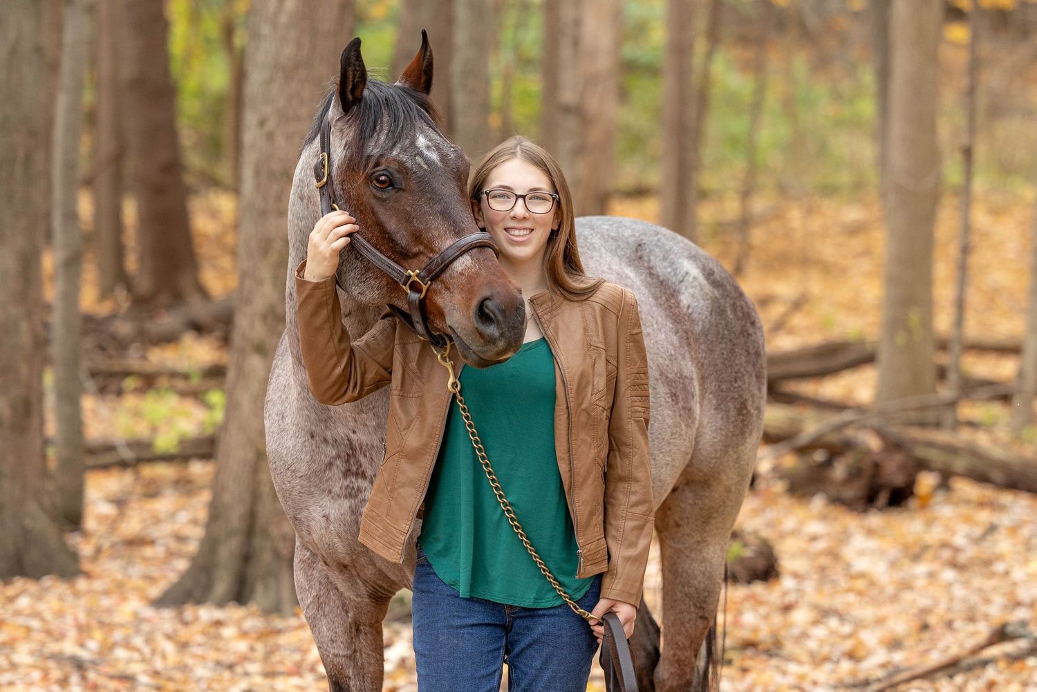 Senior photo of a girl in a green shirt and brown jacket in the woods with her bay roan horse