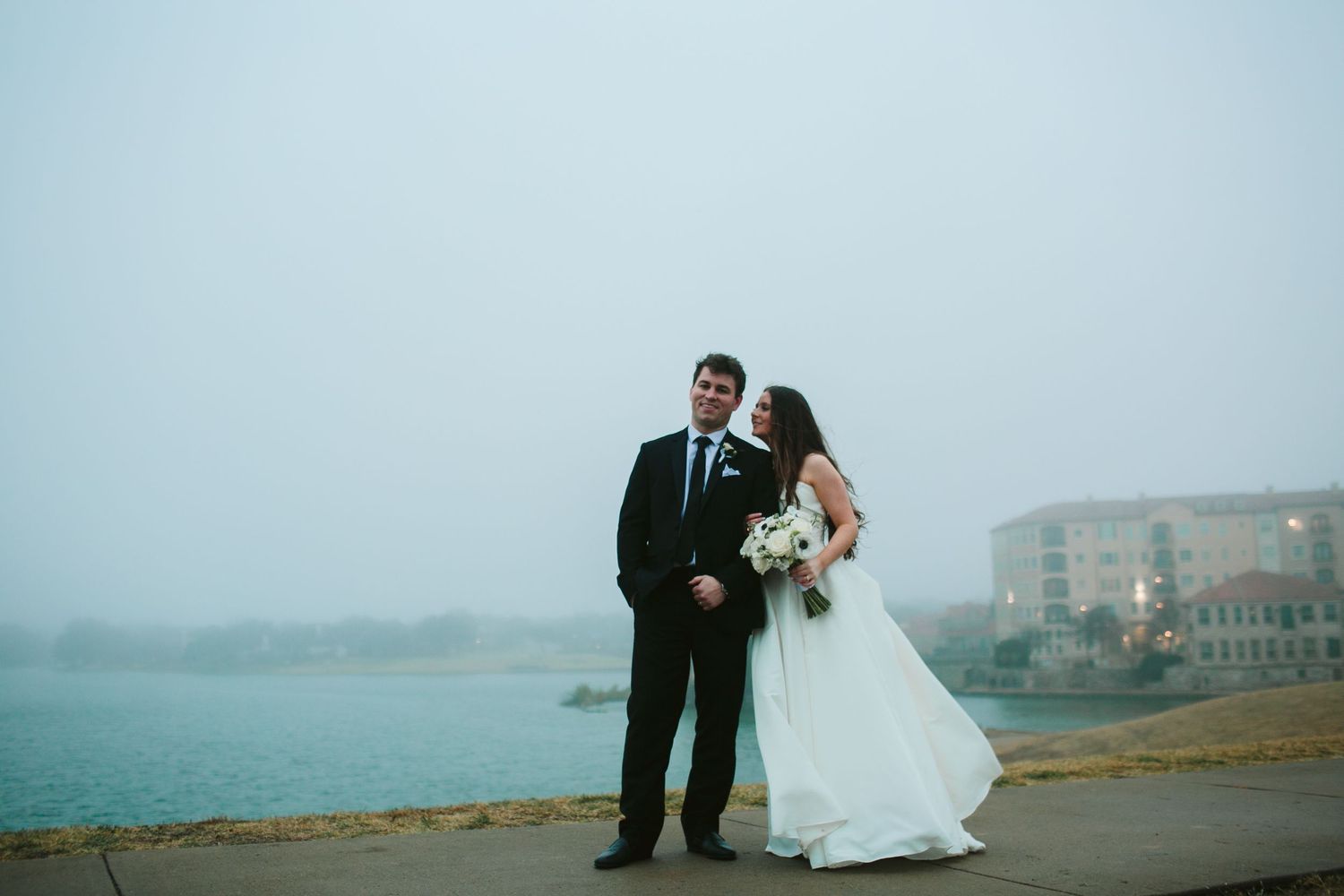 A newlywed couple poses on a foggy coastline with buildings visible in the background.