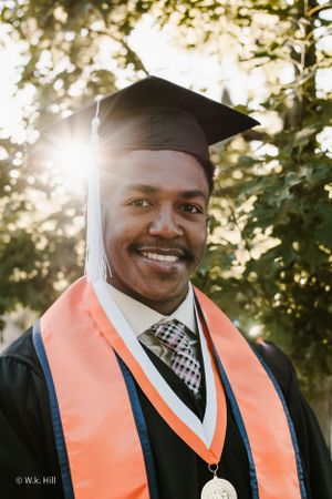 Graduate in orange stole and black cap and gown smiling in golden sunset light.