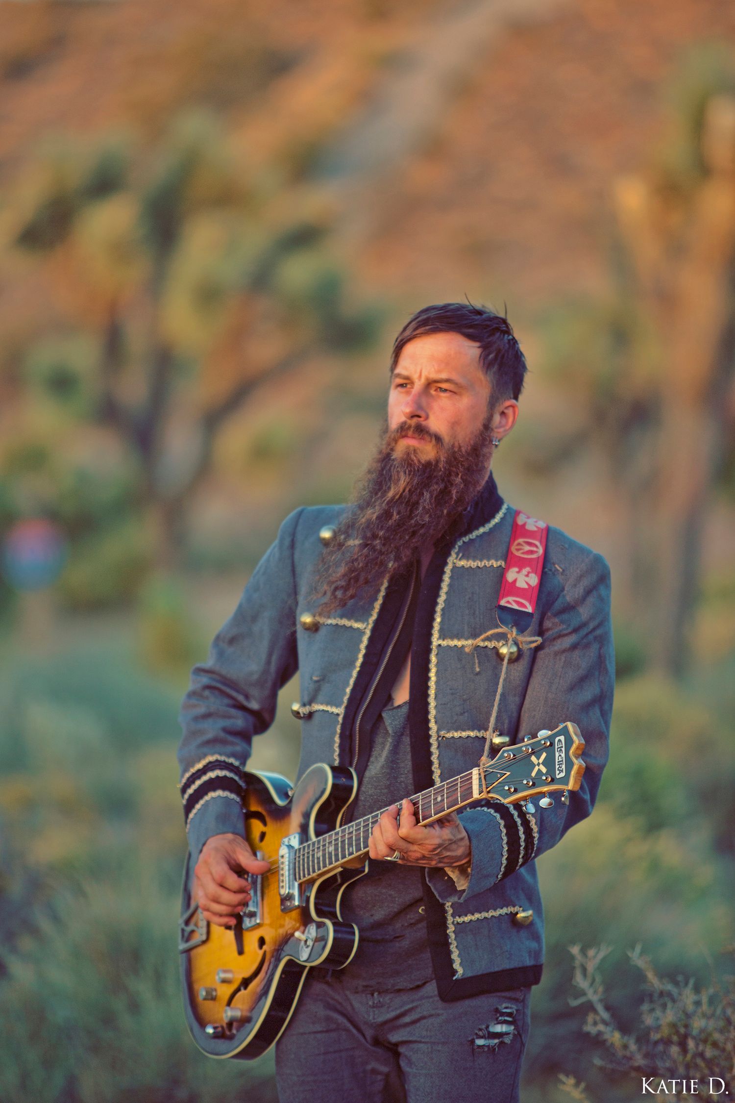 Bearded man playing guitar in desert landscape wearing decorative military-style jacket.