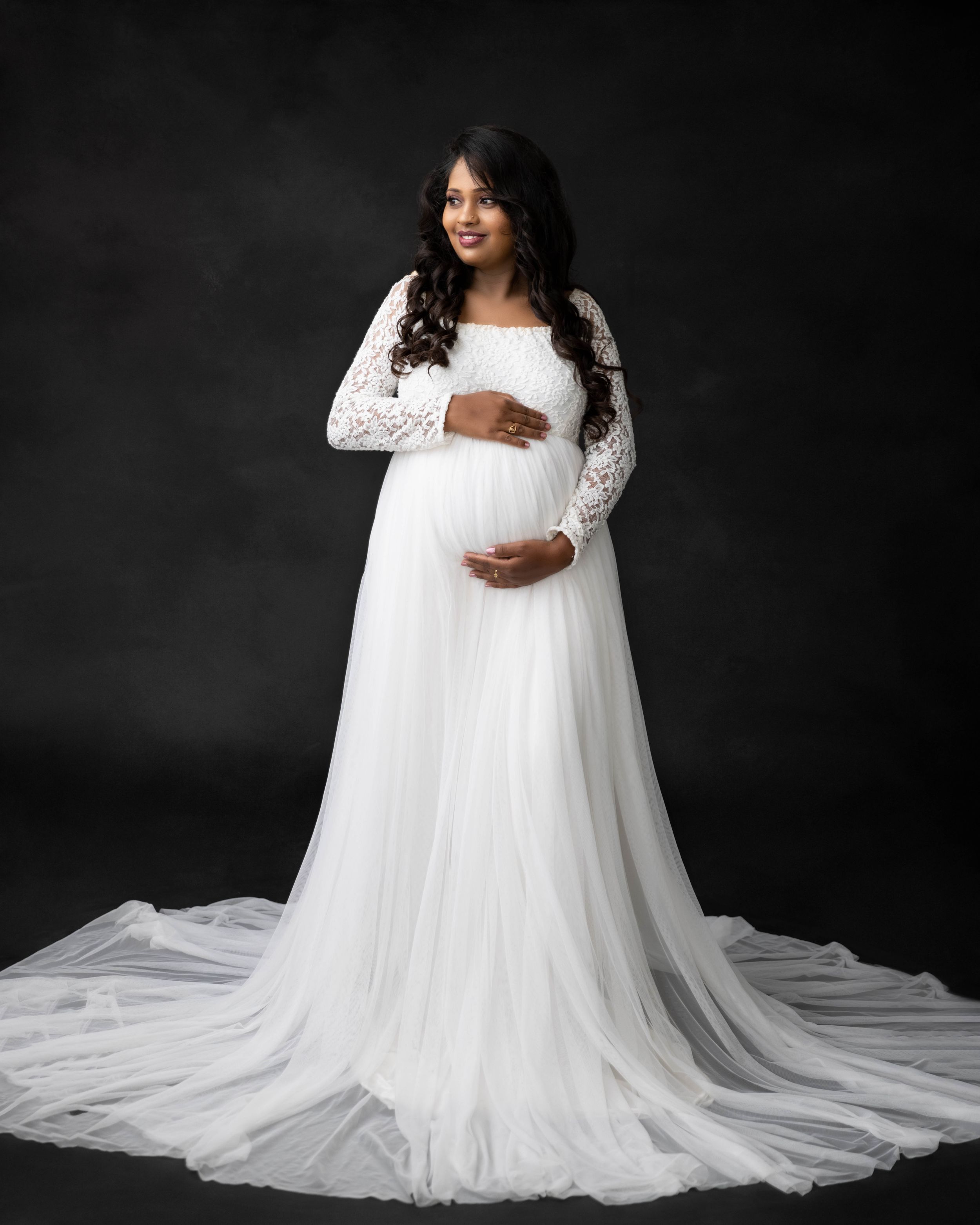 Studio maternity portrait in flowing white lace dress with long train against dark backdrop.