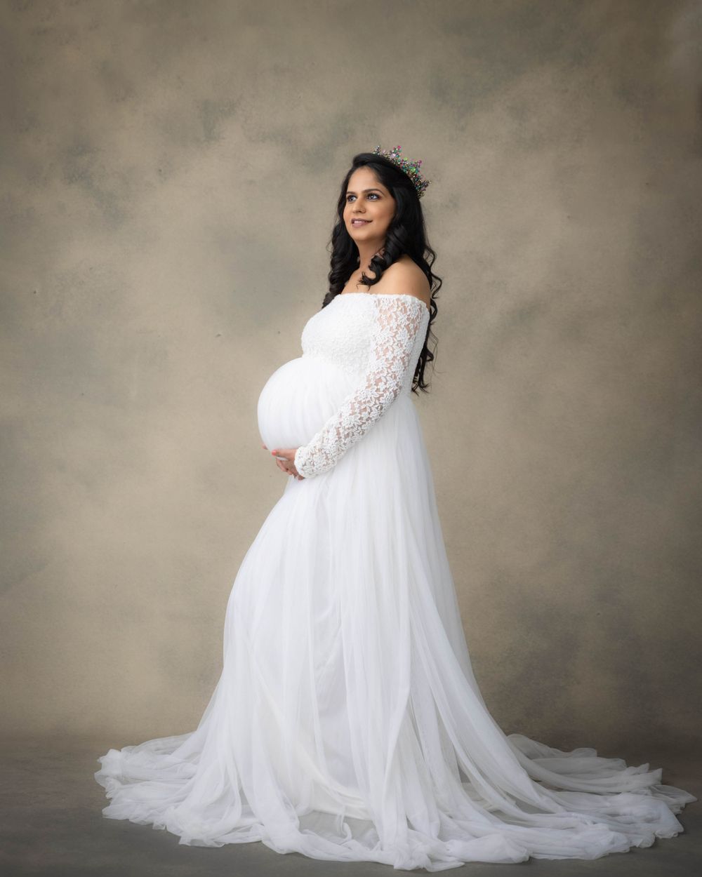 Elegant maternity portrait in white lace off-shoulder gown against neutral backdrop.
