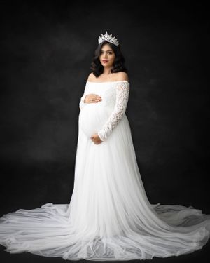 Maternity photo of woman in flowing white lace gown and crown against dark backdrop.