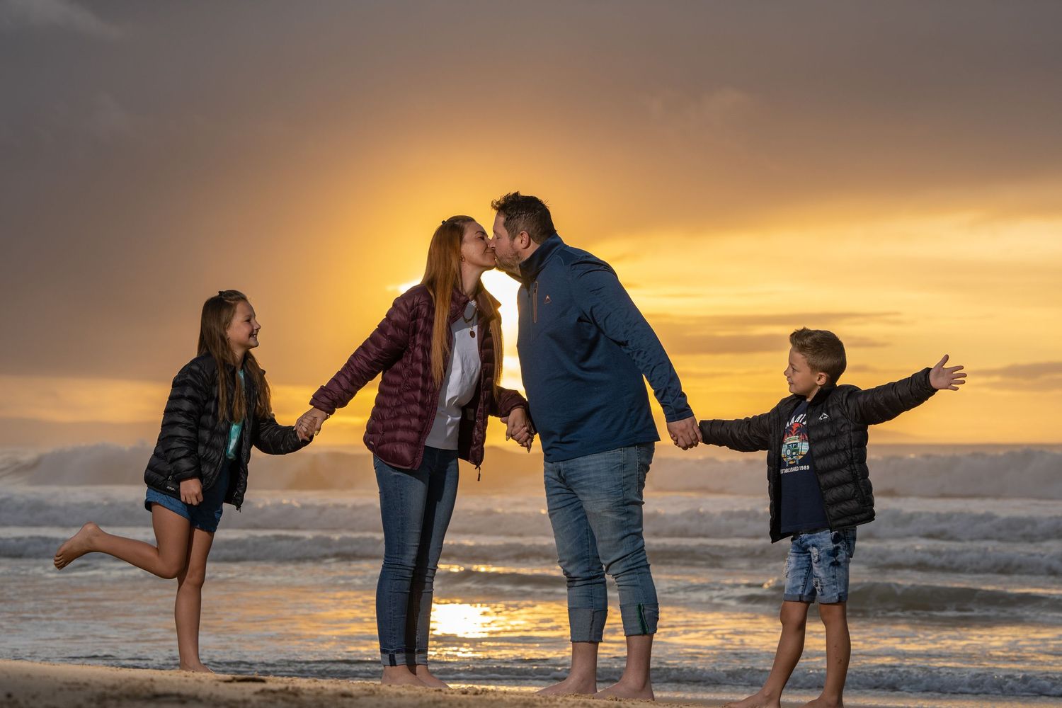 Family of four sharing a romantic sunset kiss on the beach while children hold hands.
