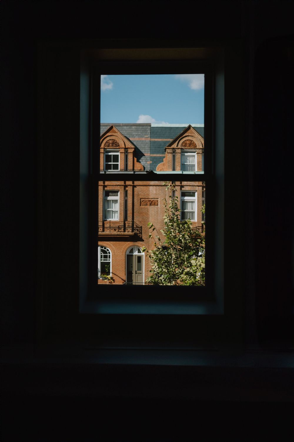 View through dark window frame of brick townhouse with dormer windows against blue sky.