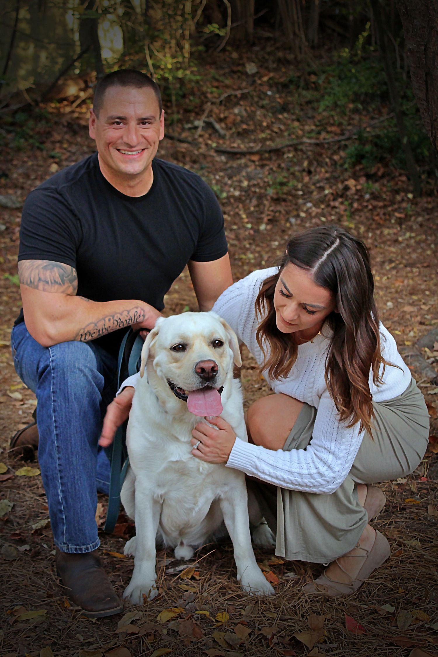 A couple poses with their happy yellow Labrador dog in an outdoor autumn setting.