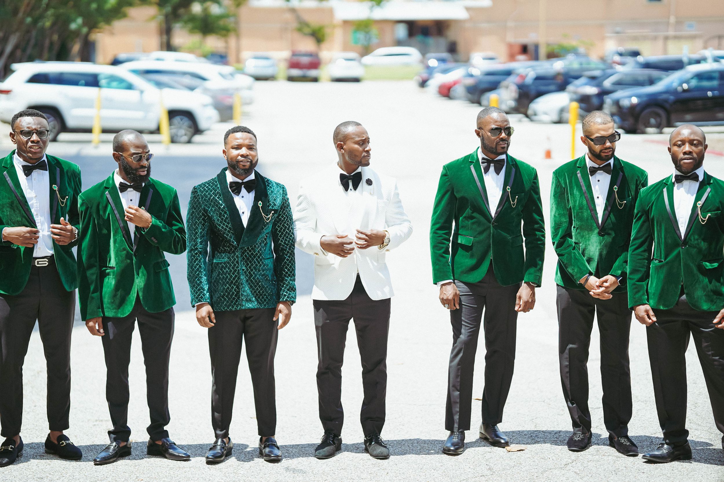 Wedding party groomsmen pose together wearing emerald green suits with one in white standing center in parking lot.