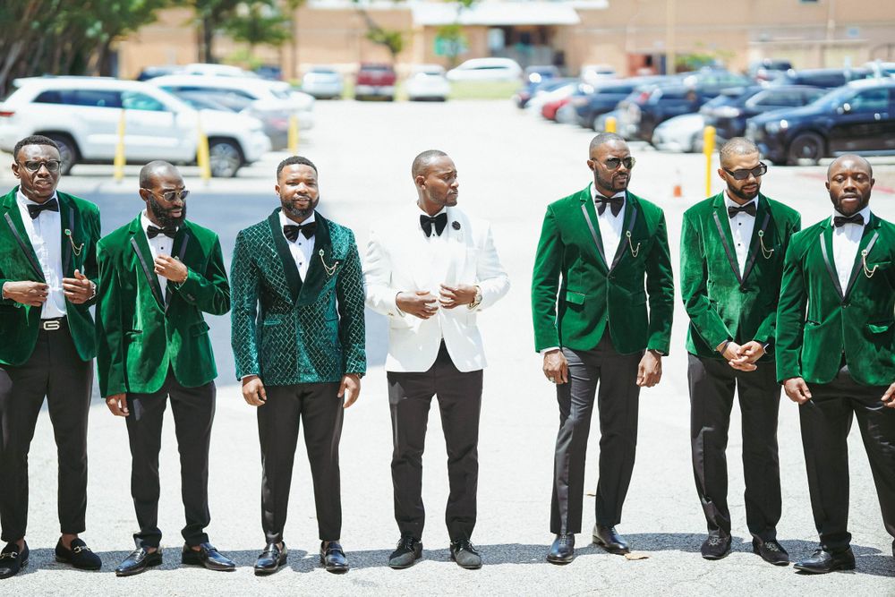 Wedding party groomsmen pose together wearing emerald green suits with one in white standing center in parking lot.