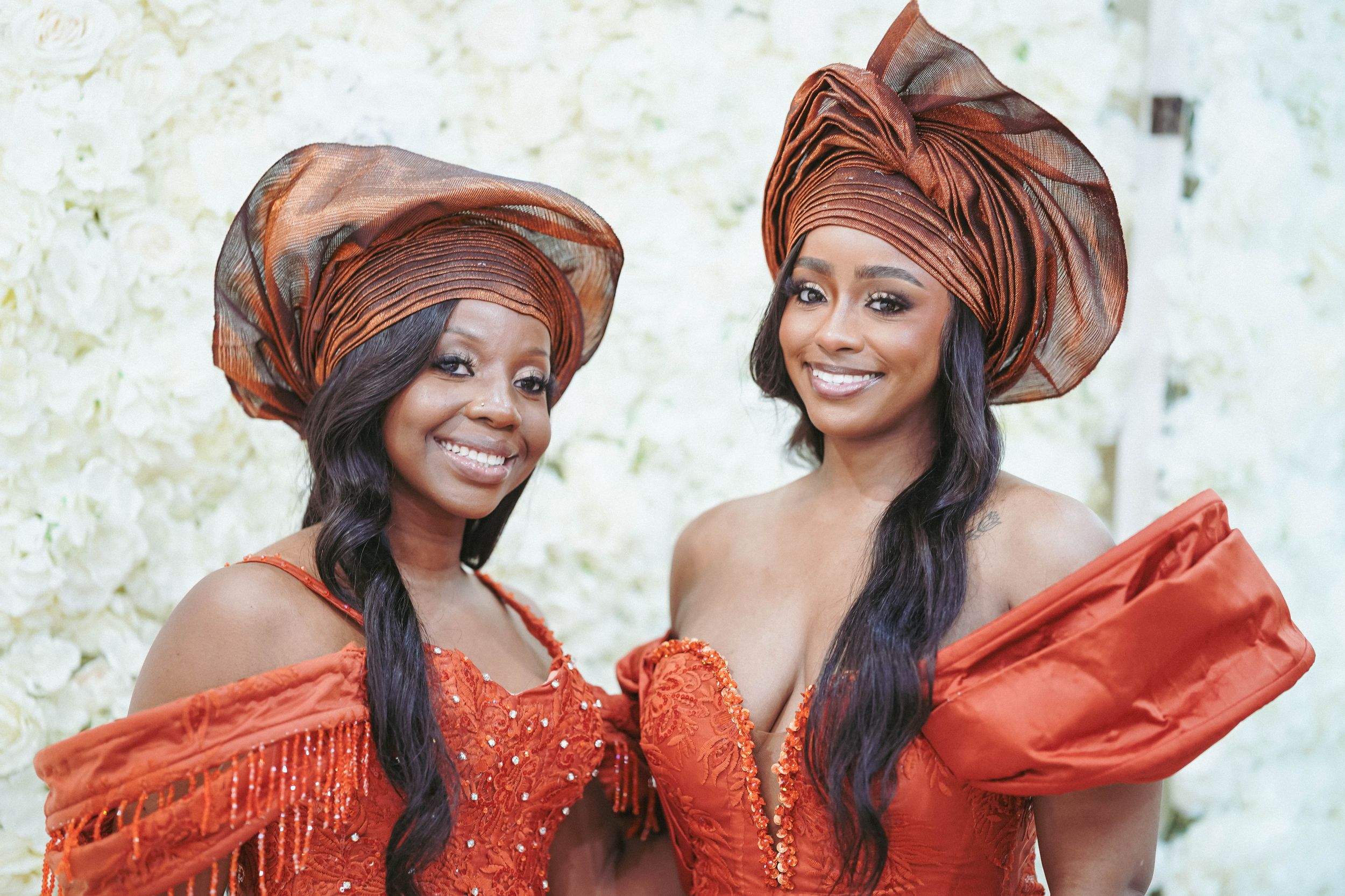 Two women in vibrant red traditional headwraps and dresses share joyful moments together.