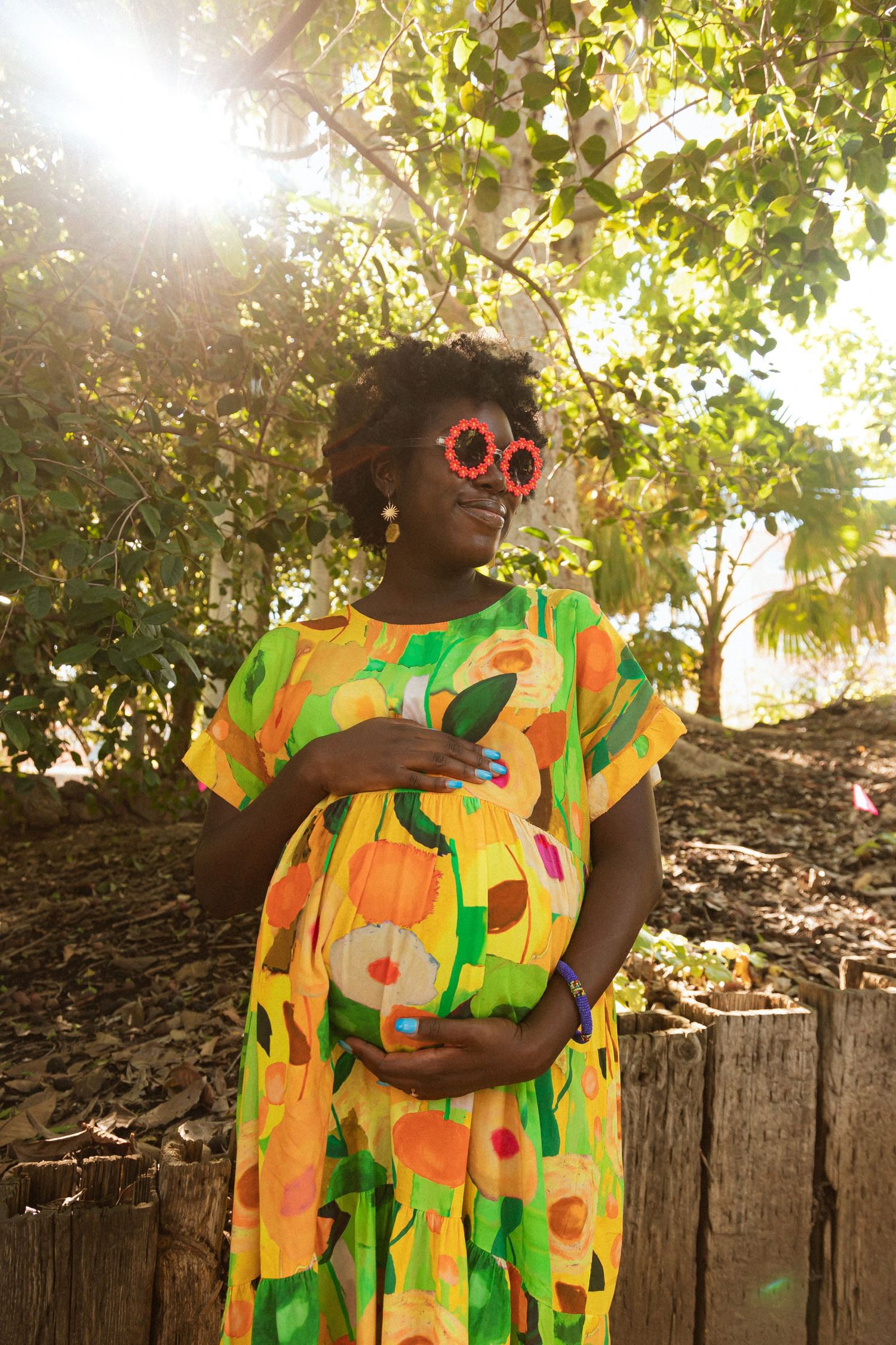 Maternity photo with person wearing vibrant floral dress and sunglasses in sunlit garden setting.