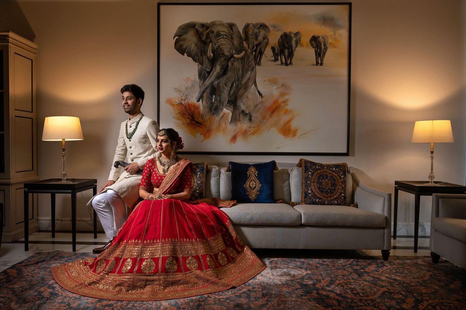 Indian bride in red lehenga and groom in white sit elegantly before elephant artwork in a luxurious hotel suite.