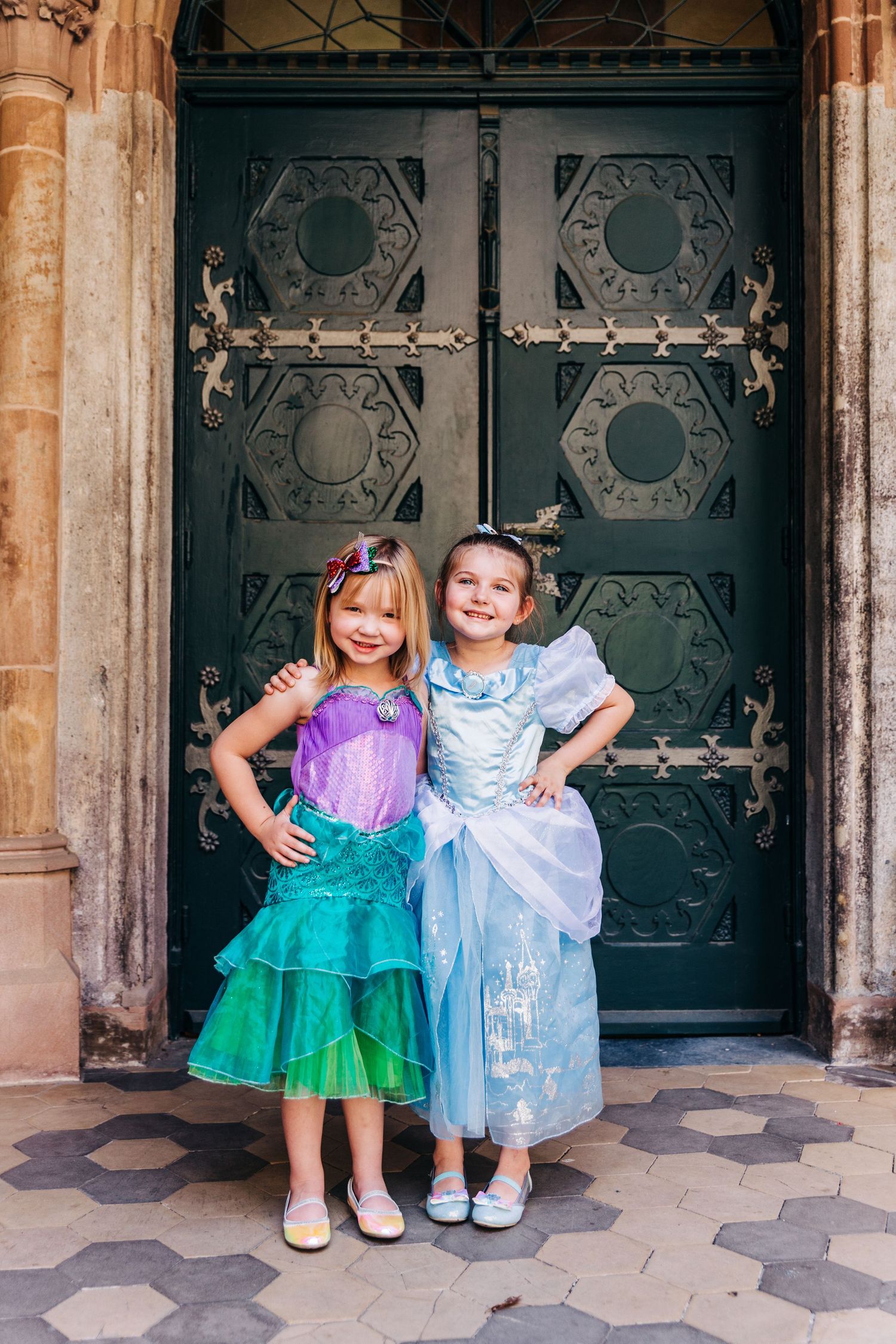 Two friends in princess costumes pose together in front of an ornate Gothic door.
