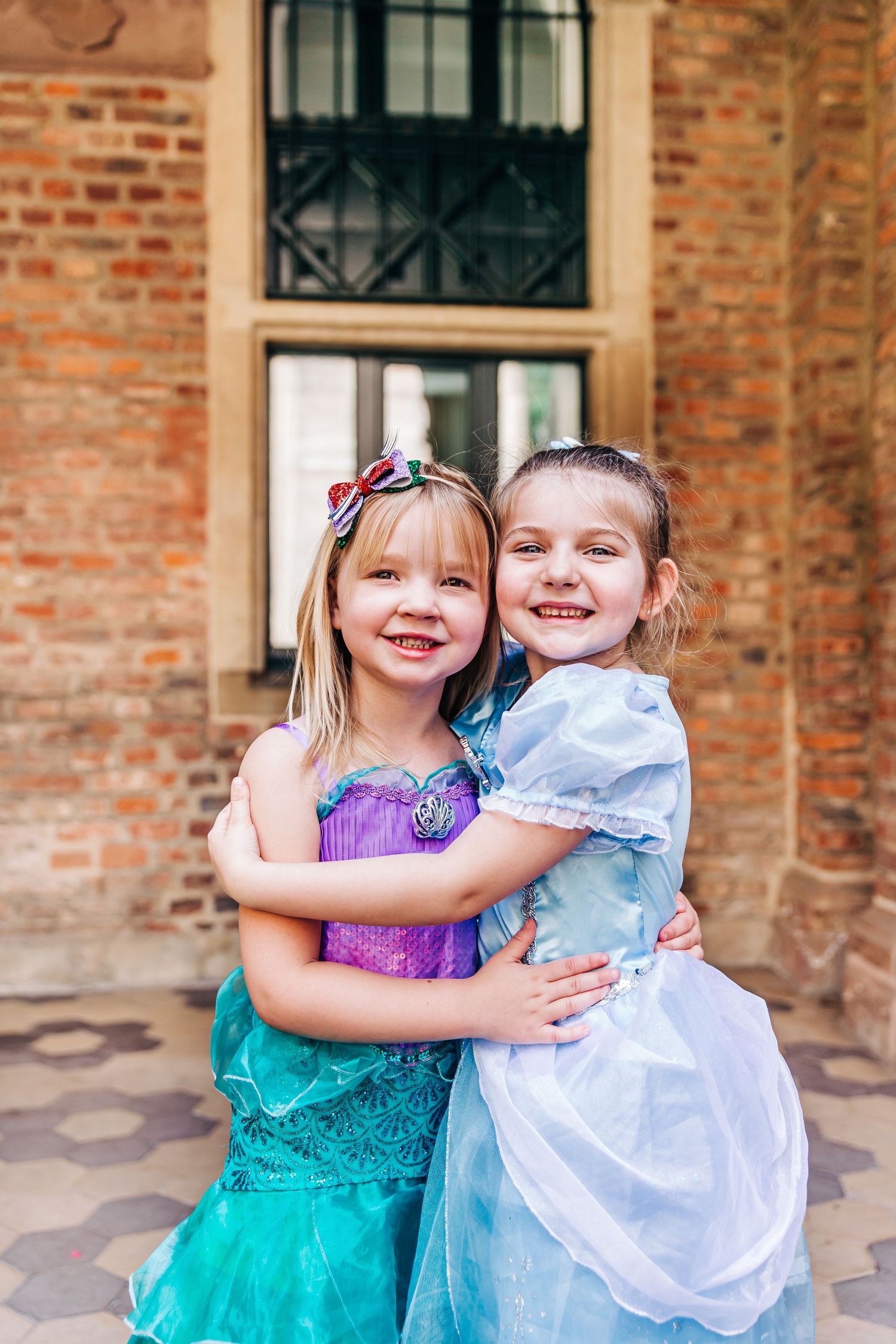 Friends in princess costumes share a warm embrace in front of brick architecture.