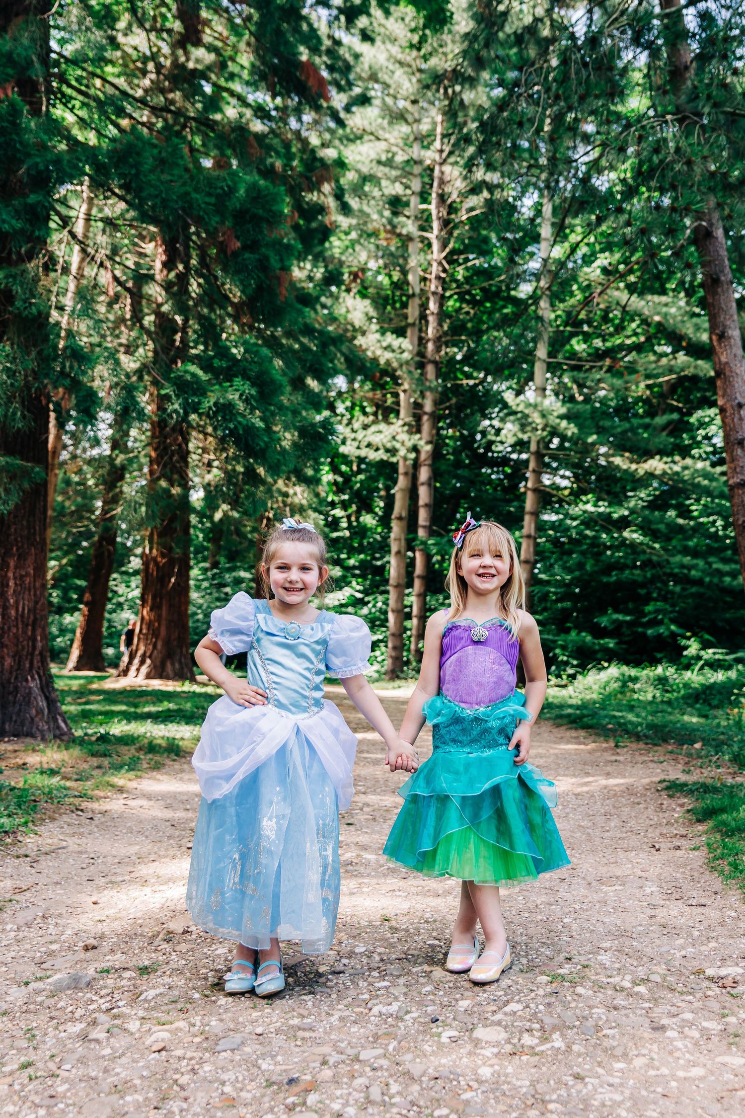 Two young fairy tale princesses in blue and green dresses pose together on a forest path.