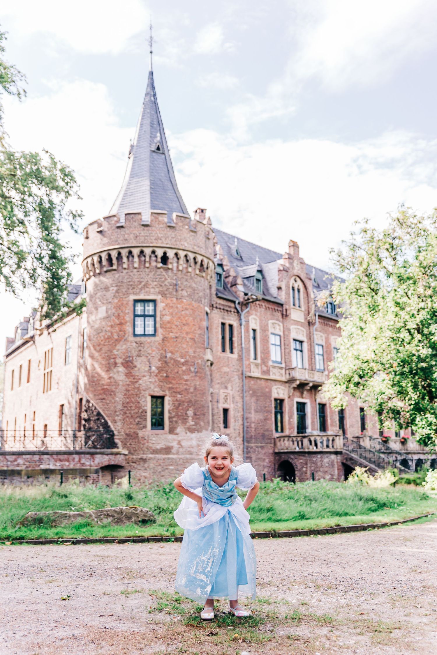 A princess in a blue dress poses in front of a magnificent fairytale castle with stone towers.